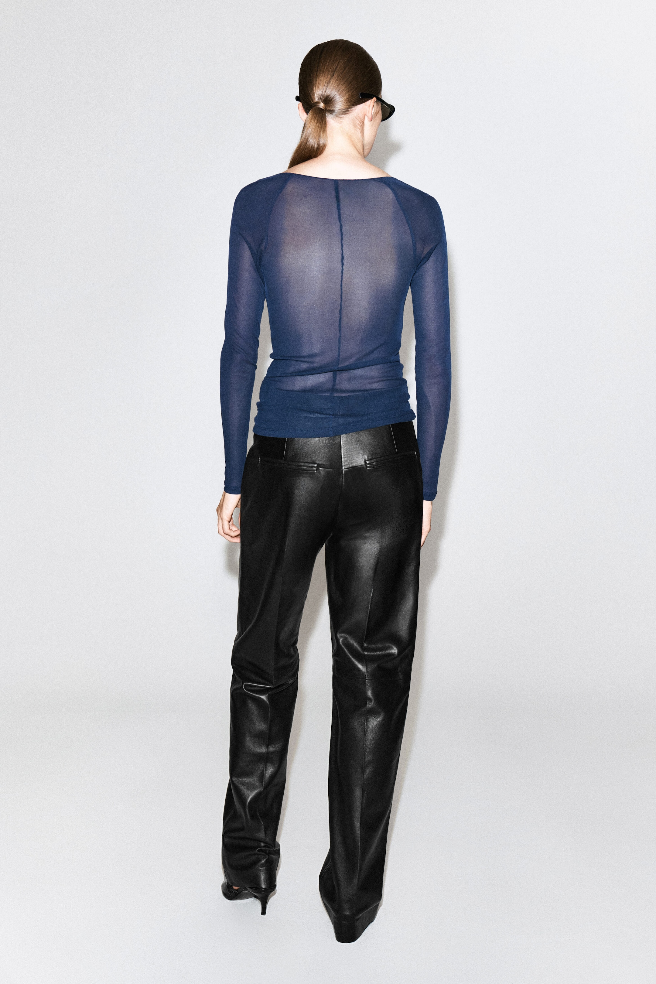 Le mannequin porte un haut fin en soie côtelée bleu marine transparent, mettant en valeur ses manches longues, sa coupe ajustée et une couture verticale qui descend le long du dos. Le haut léger est agencé avec un pantalon en cuir noir à taille haute.