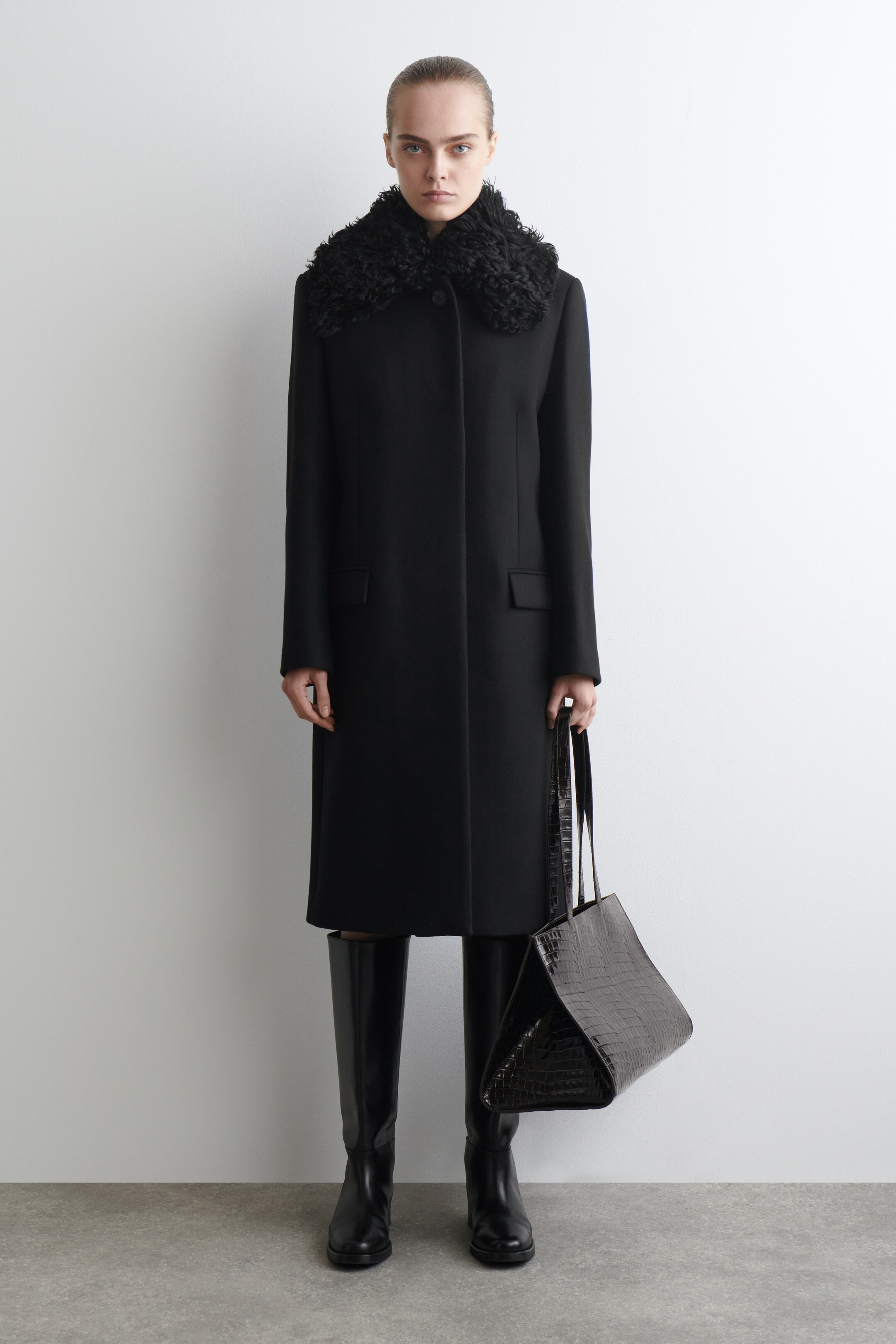 View larger image: A black coat is facing forward, featuring a prominent black faux fur collar. It is displaying a straight, midi-length silhouette with long sleeves and two flap pockets on the front.