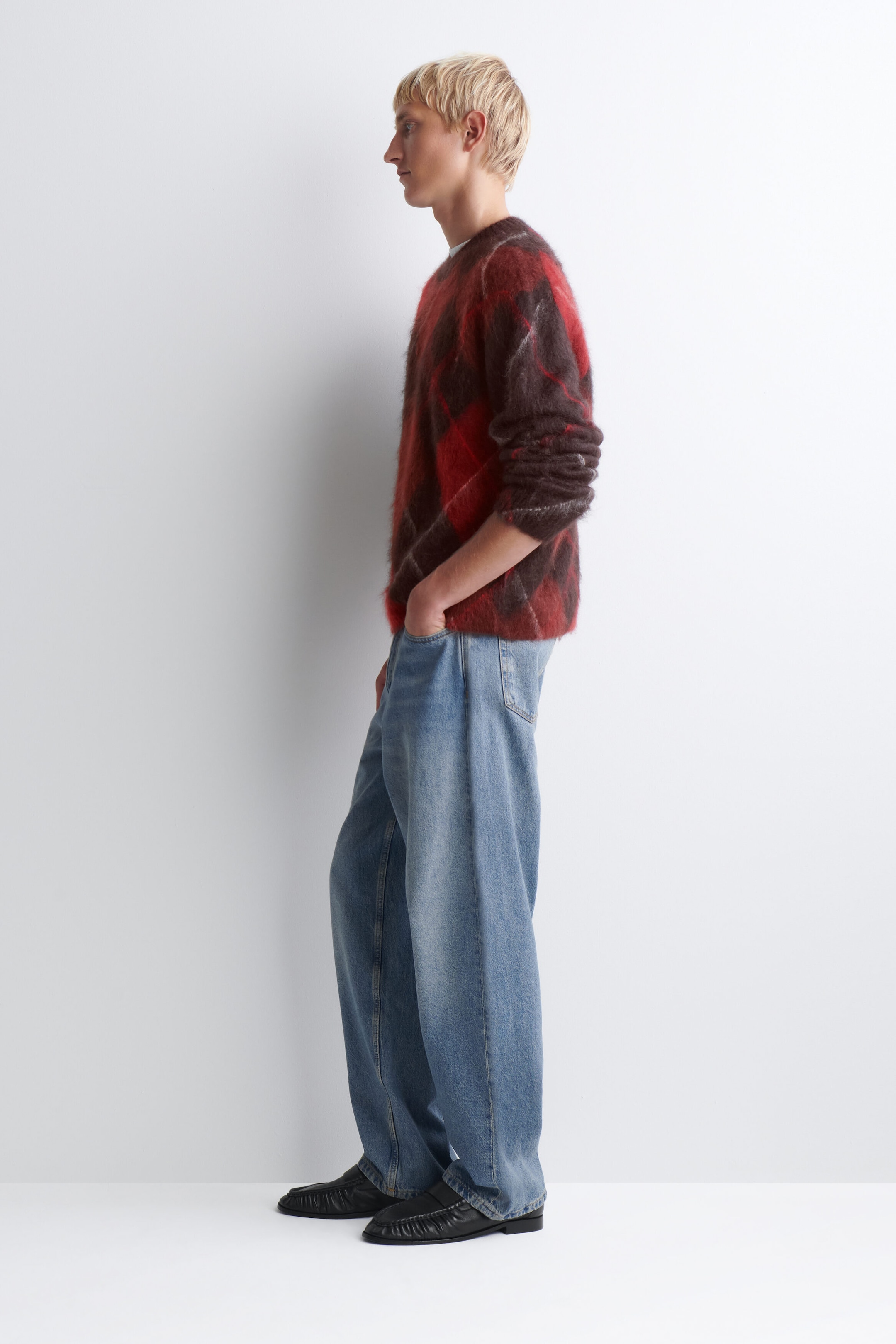 Ein flauschiger Rundhalspullover mit rotem und dunkelbraunem Argyle-Muster wird mit locker sitzenden, weit geschnittenen hellblauen Jeans kombiniert. Das Outfit wird in einer seitlichen Profilansicht nach links gezeigt, wodurch die lockere Passform und die Textur beider Teile hervorgehoben werden.