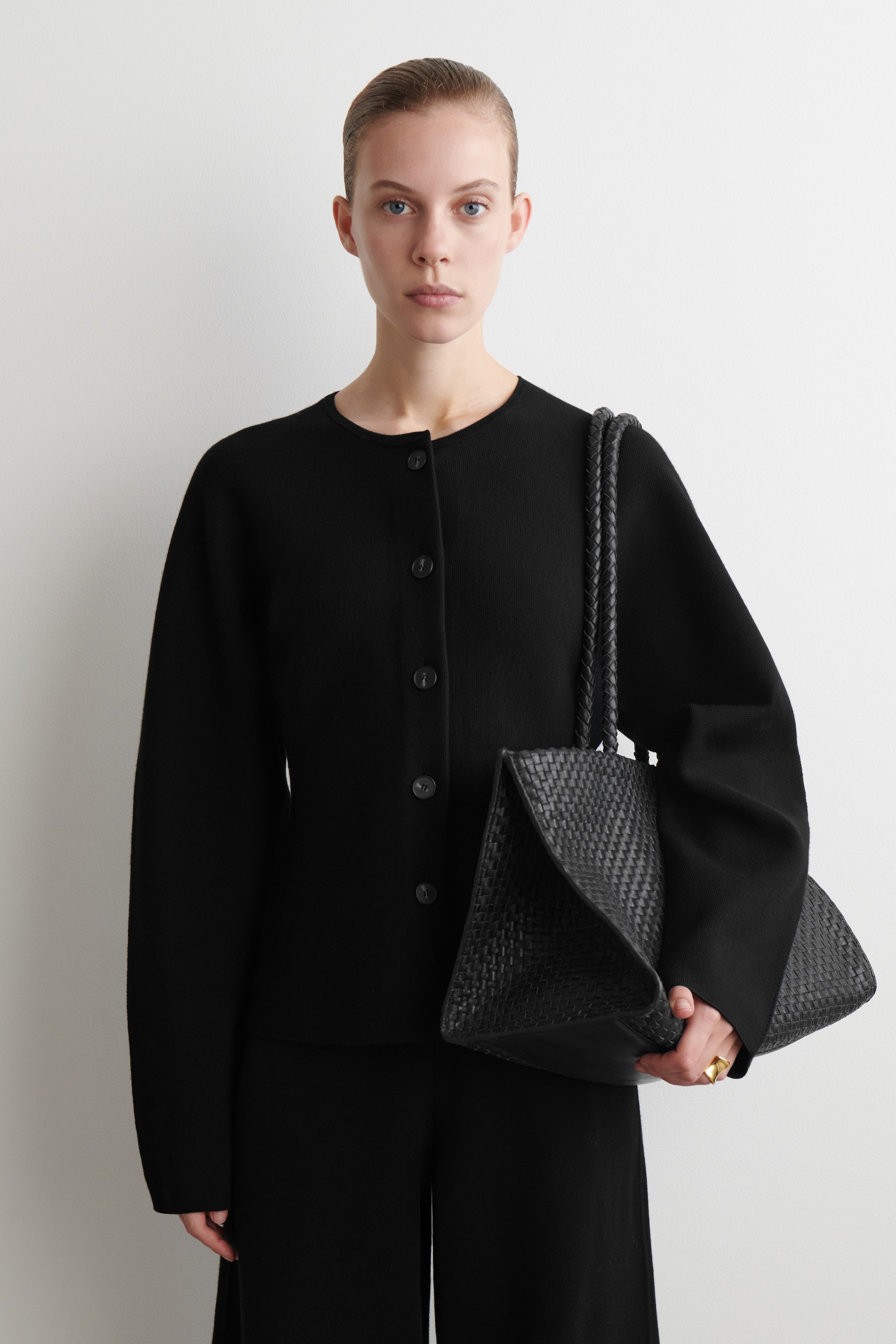 Agrandir l'image: Un cardigan en tricot noir, avec un col rond et une fermeture à boutons sur le devant, est porté avec un pantalon noir. Un sac cabas en cuir tressé noir avec des poignées tressées est porté, sa surface texturée étant visiblement inclinée sur le côté.