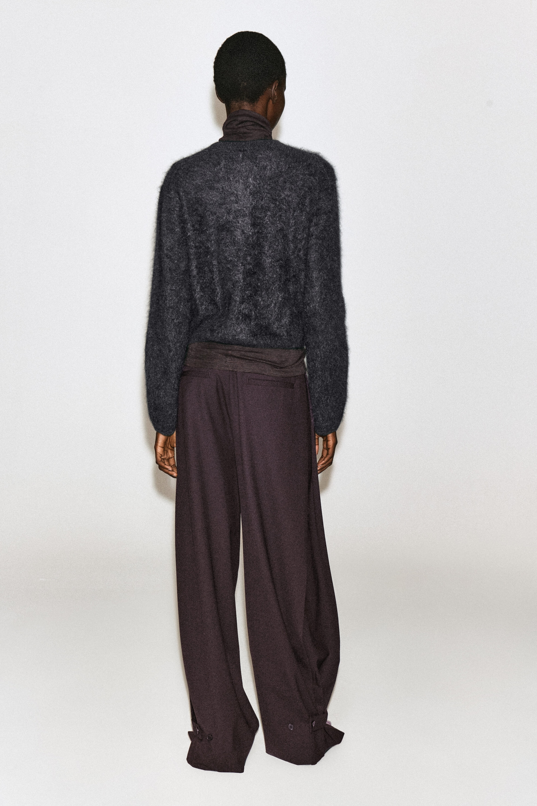 A dark grey brushed cashmere crew-neck cardigan is being worn by the model, showing its textured, fuzzy finish from the back. The cardigan is layered over a dark brown turtleneck and styled with loose, dark wide-leg trousers. A dark grey brushed cashmere crew-neck cardigan is being worn by the model, showing its textured, fuzzy finish from the back. The cardigan is layered over a dark brown turtleneck and styled with loose, dark wide-leg trousers.