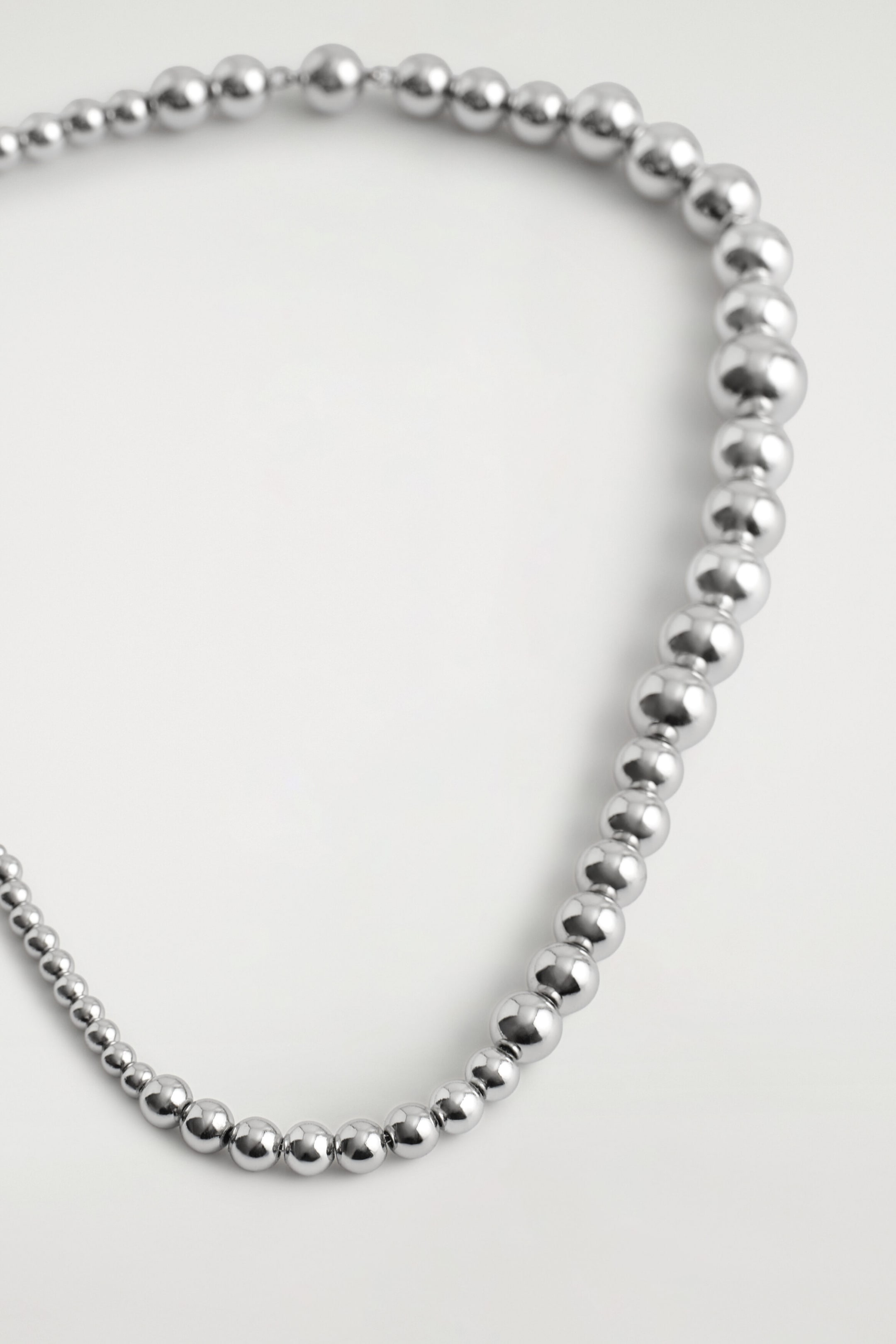 Un collier en perles présente des perles sphériques polies, de teinte argentée, de tailles graduées disposées en boucle courbe. Les perles métalliques hautement réfléchissantes passent de petites à grandes le long du rang. Un collier en perles présente des perles sphériques polies, de teinte argentée, de tailles graduées disposées en boucle courbe. Les perles métalliques hautement réfléchissantes passent de petites à grandes le long du rang.