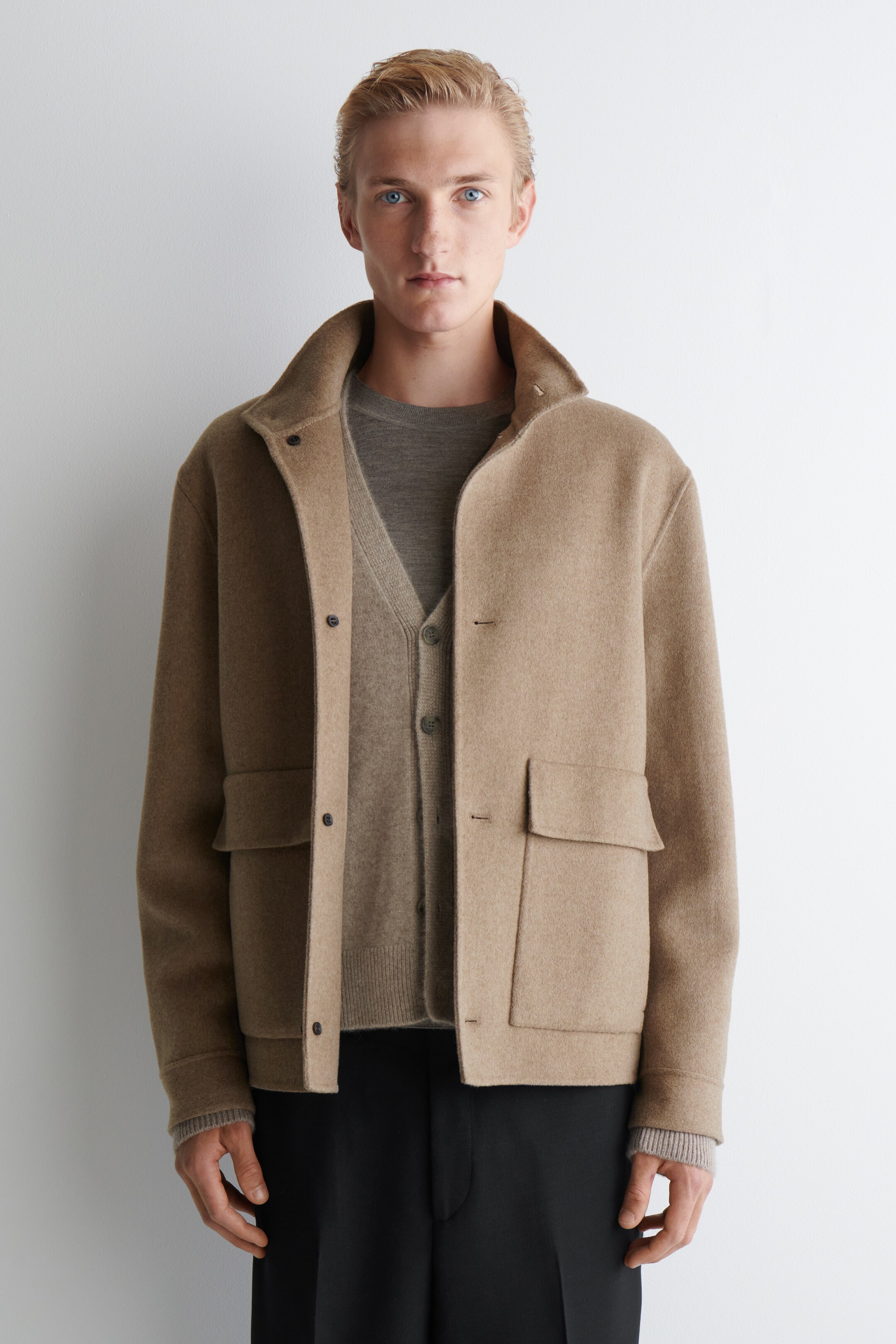 Ver imagen más grande: Una chaqueta corta de camello está siendo usada por la modelo, con un cuello alto, una tapeta de botones a presión y dos grandes bolsillos con solapa. Está superpuesta sobre un cárdigan de punto tonal y un suéter de cuello redondo.