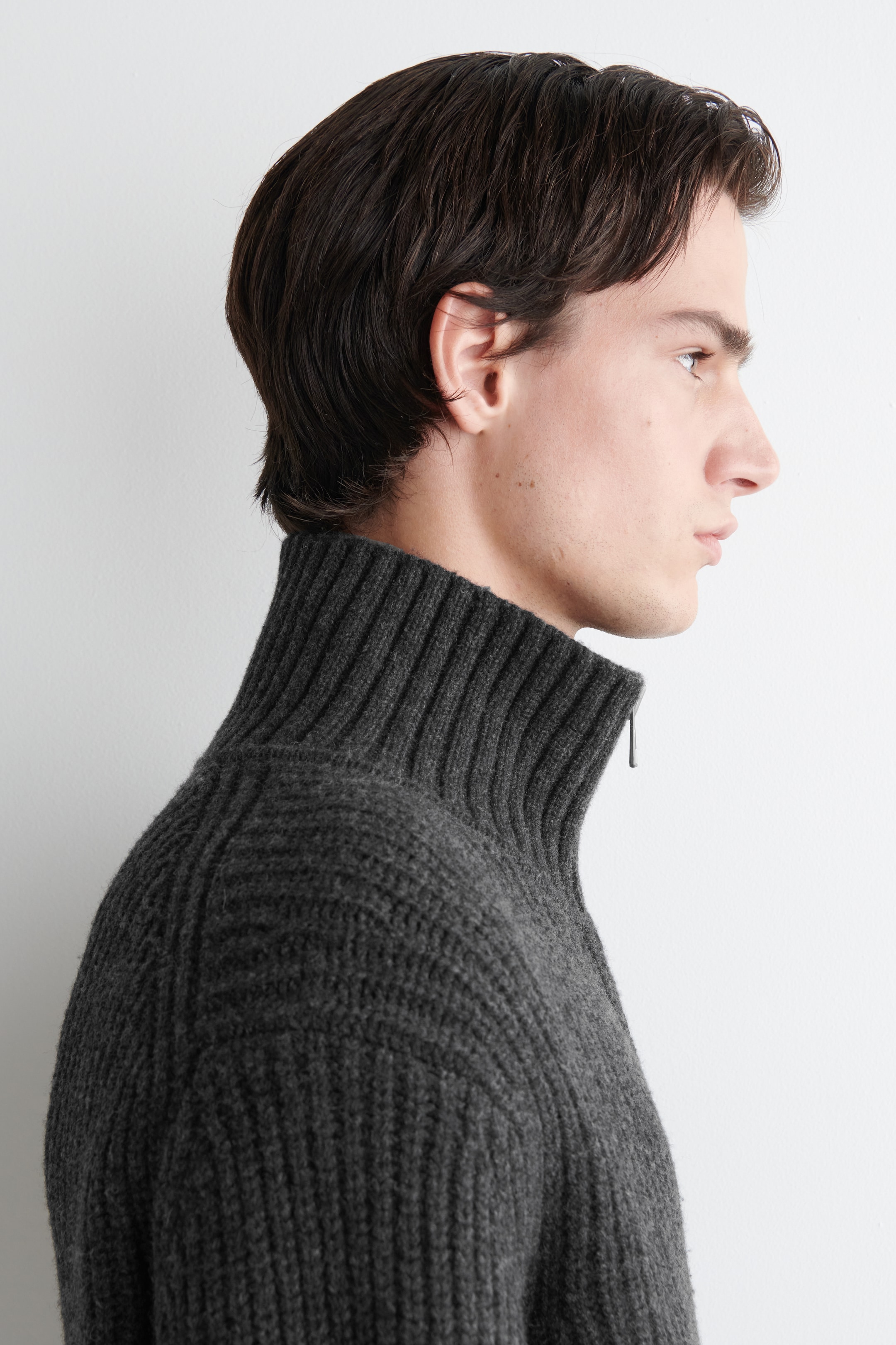 Agrandir l'image: Un pull gris foncé en maille côtelée est présenté de profil. Il est doté d'un col montant côtelé avec une fermeture éclair partiellement ouverte à l'avant, et la texture côtelée prononcée est visible sur l'épaule et la partie supérieure du corps.