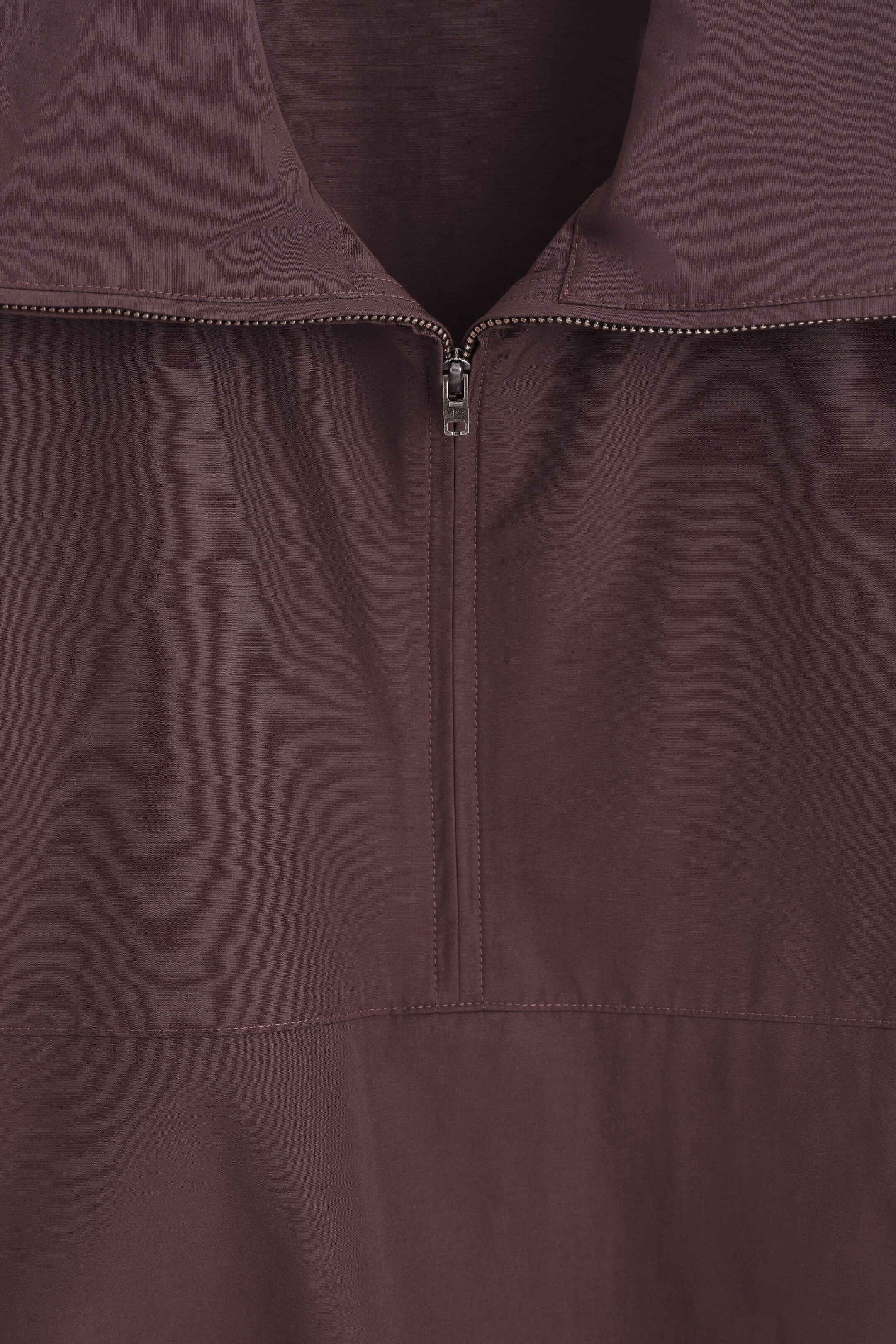 A close-up view shows a dark plum-coloured garment featuring a silver-tone half-zip closure, a wide collar, and topstitched seam details. A close-up view shows a dark plum-coloured garment featuring a silver-tone half-zip closure, a wide collar, and topstitched seam details.