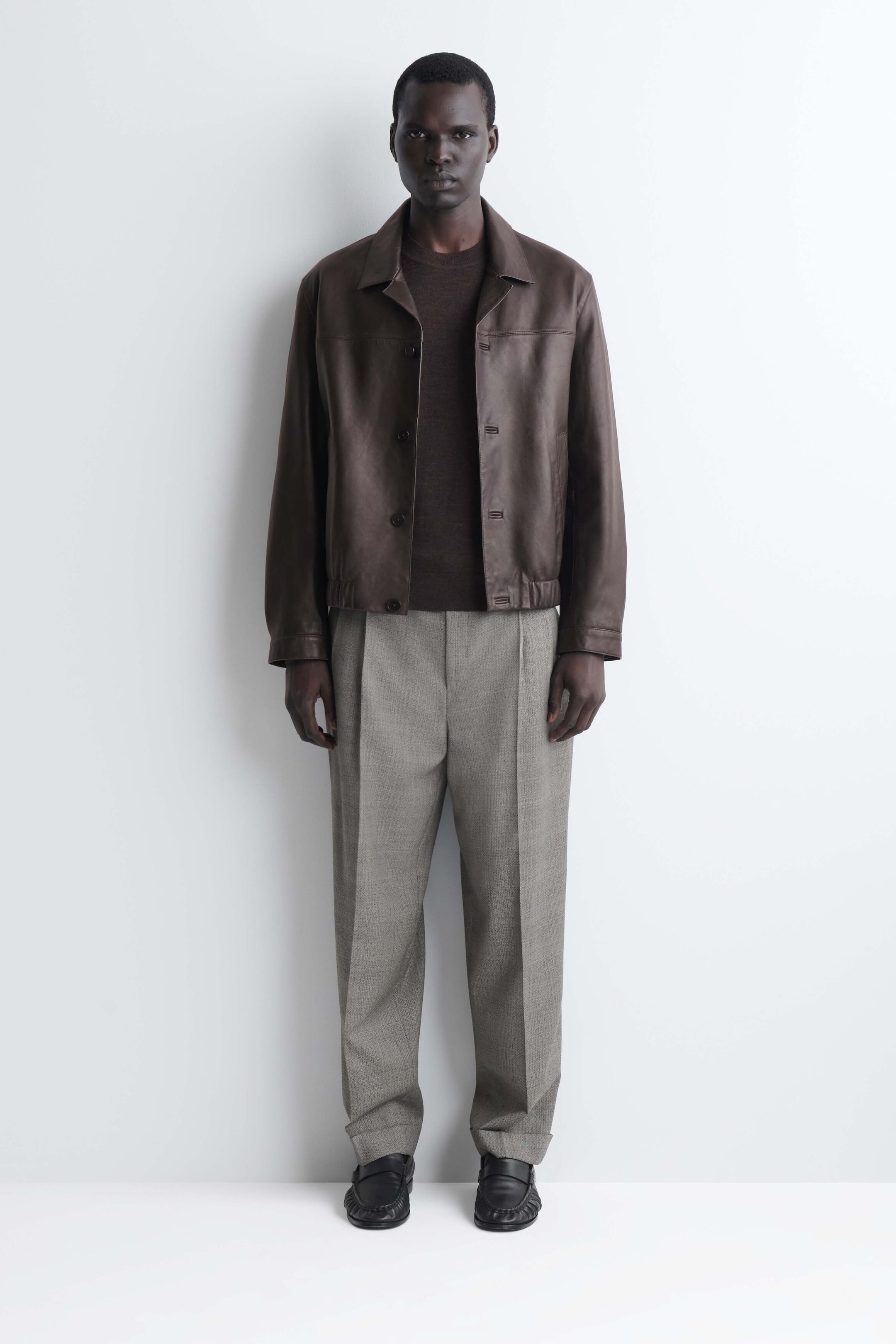The slim merino wool crew-neck sweater in dark brown is shown from the front, layered underneath an open brown leather jacket. The fine-knit sweater shows a close fit and a classic crew neckline, paired with light gray trousers. The slim merino wool crew-neck sweater in dark brown is shown from the front, layered underneath an open brown leather jacket. The fine-knit sweater shows a close fit and a classic crew neckline, paired with light gray trousers.
