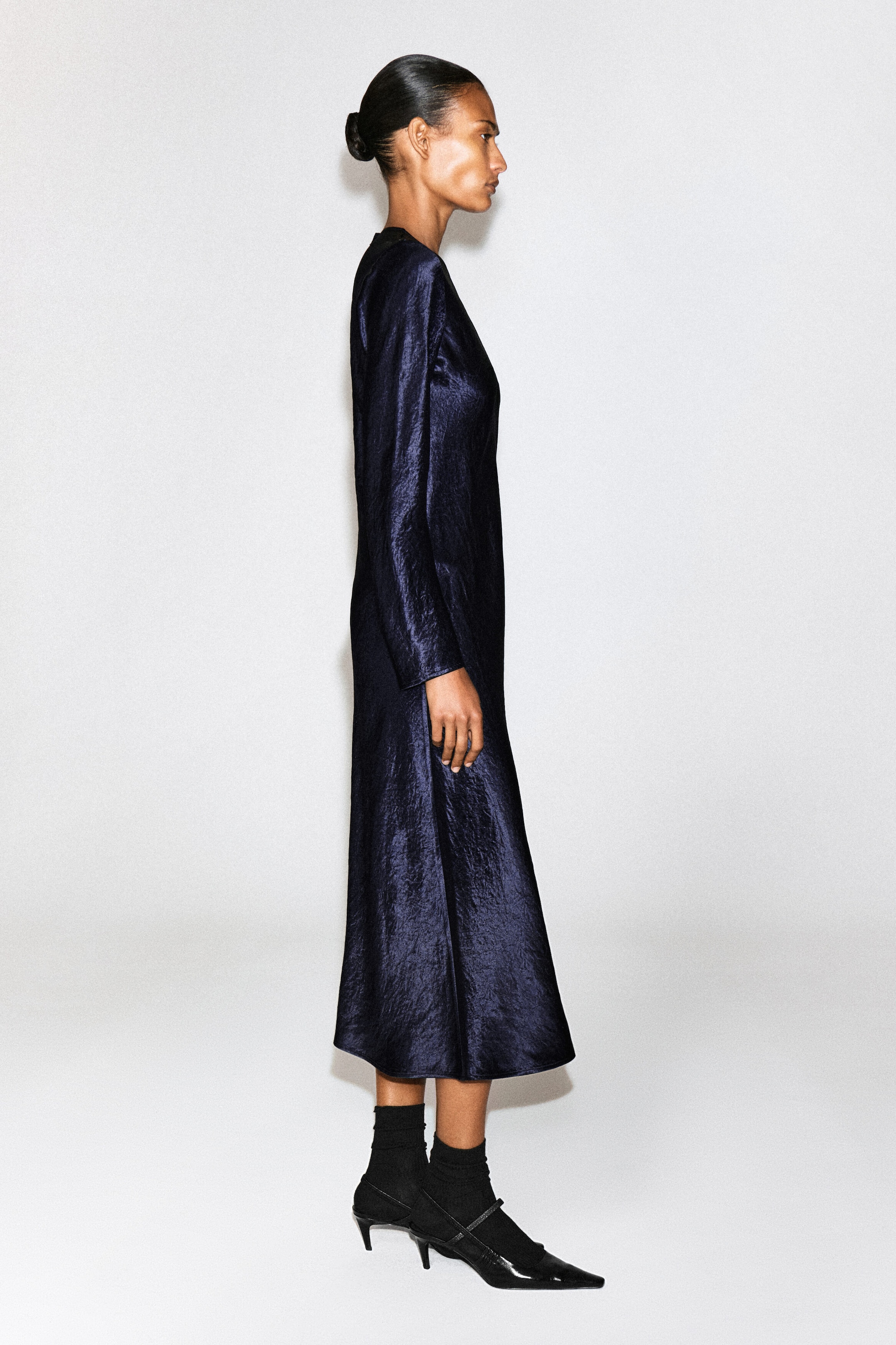 A navy blue textured satin long-sleeve maxi dress is worn by the model in side profile. The dress features a high neckline and a crinkled texture, styled with black socks and black pointed-toe kitten heels. A navy blue textured satin long-sleeve maxi dress is worn by the model in side profile. The dress features a high neckline and a crinkled texture, styled with black socks and black pointed-toe kitten heels.