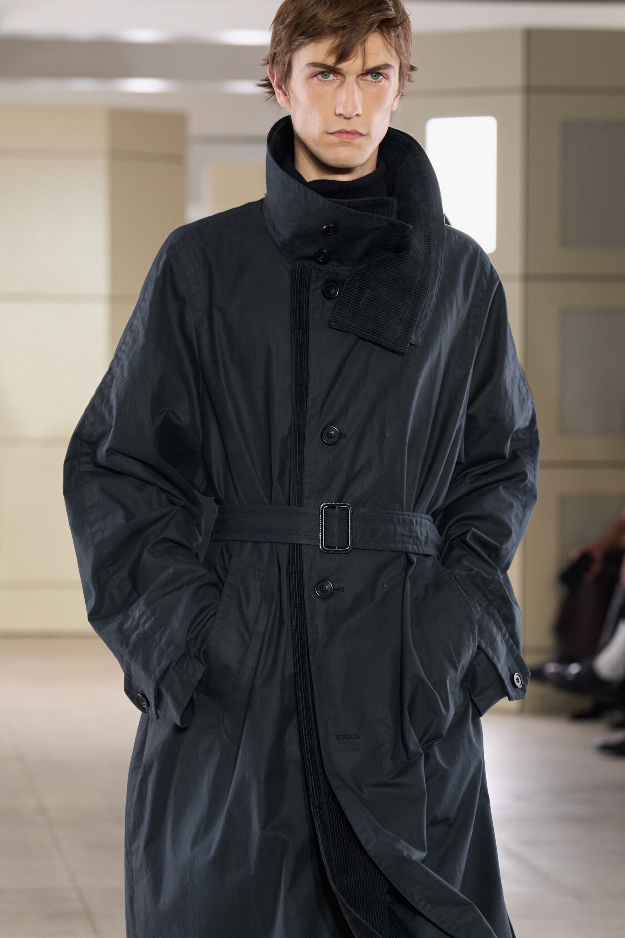 A dark navy waxed cotton trench coat is shown from the front, featuring a high, asymmetrical collar lined with matching corduroy fabric. The coat is cinched at the waist with a matching fabric belt and layered over a black high-neck top. A dark navy waxed cotton trench coat is shown from the front, featuring a high, asymmetrical collar lined with matching corduroy fabric. The coat is cinched at the waist with a matching fabric belt and layered over a black high-neck top.