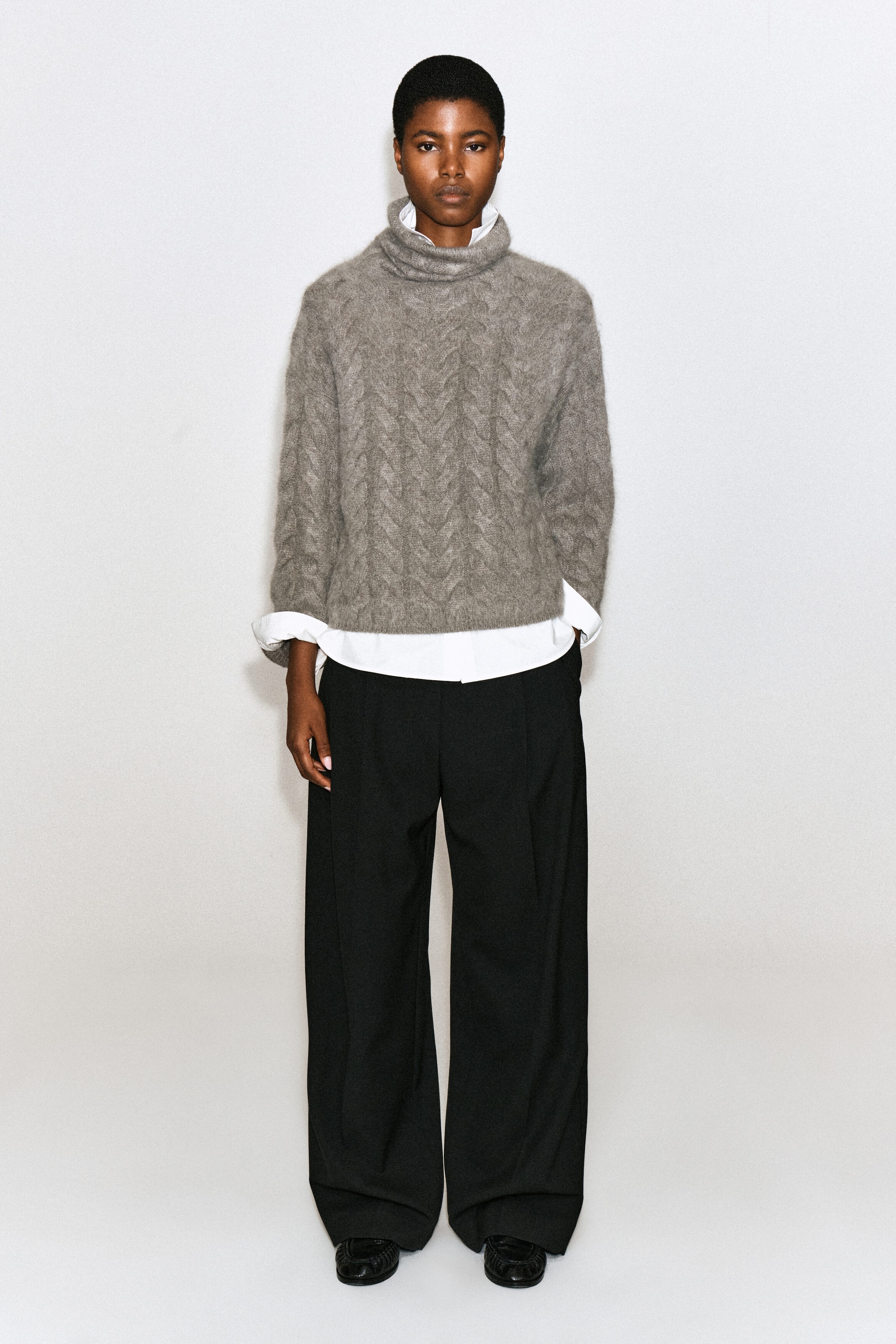 Un chandail gris en cachemire brossé à col cheminée et maille torsadée est porté par le mannequin, superposé à une chemise blanche à col et assorti à un pantalon noir à jambe large. Le chandail présente un motif torsadé texturé et un col cheminée décontracté, avec le col, les poignets retroussés et l’ourlet de la chemise blanche bien visibles. Un chandail gris en cachemire brossé à col cheminée et maille torsadée est porté par le mannequin, superposé à une chemise blanche à col et assorti à un pantalon noir à jambe large. Le chandail présente un motif torsadé texturé et un col cheminée décontracté, avec le col, les poignets retroussés et l’ourlet de la chemise blanche bien visibles.