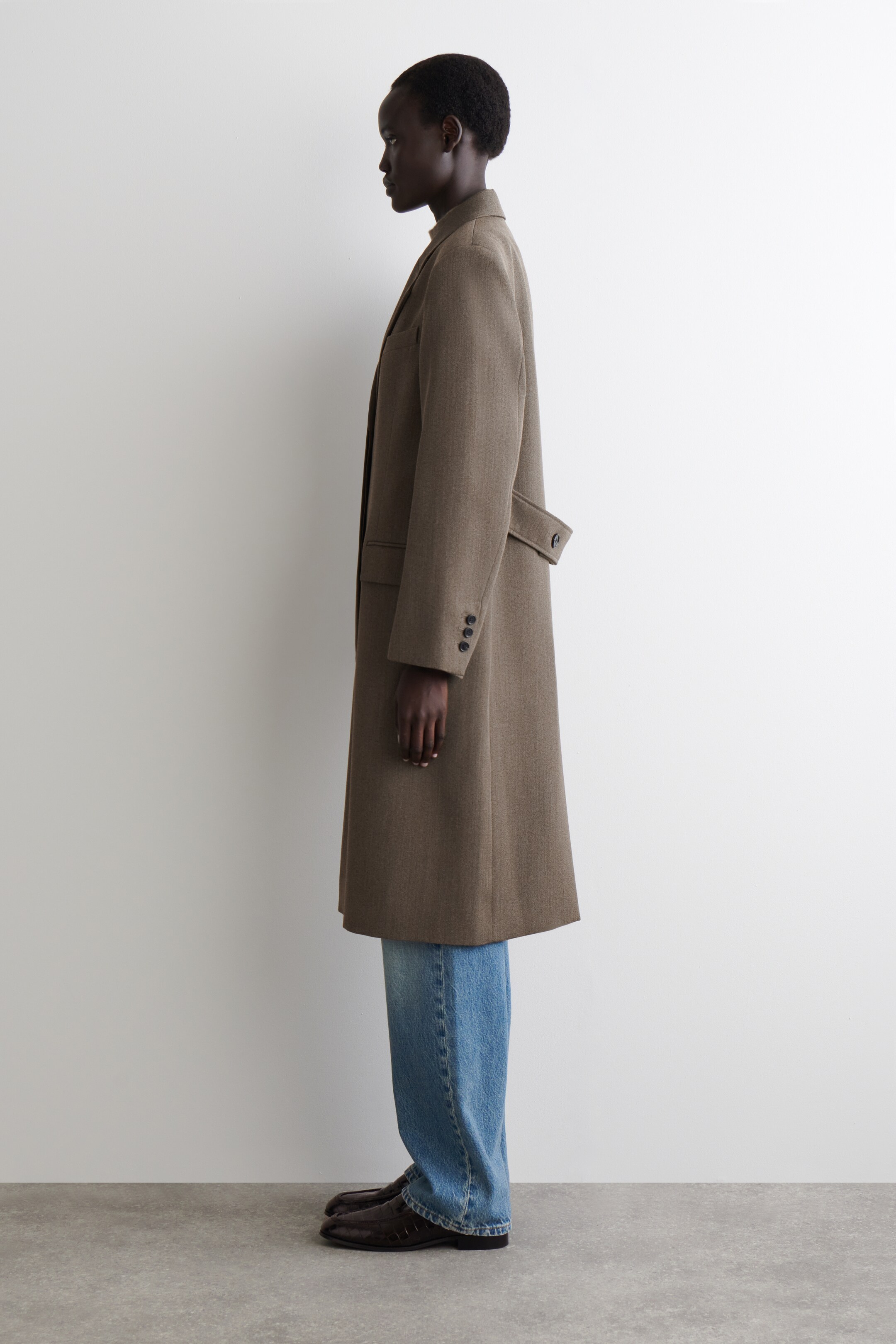 Agrandir l'image: Un long manteau texturé marron est présenté de côté, avec une coupe droite et décontractée qui s'étend jusqu'au milieu du mollet. Il présente de longues manches avec trois boutons sur les poignets, une poche passepoilée horizontale et une demi-ceinture avec un bouton à l'arrière.