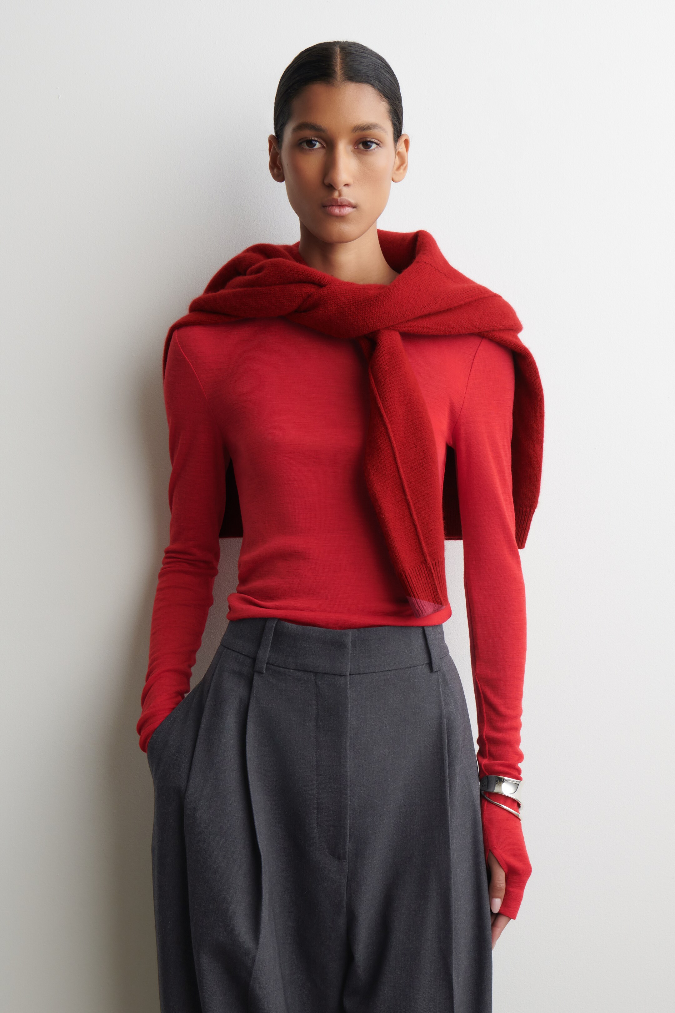 Agrandir l'image: Un haut rouge ajusté à manches longues, avec des poignets allongés et des trous pour les pouces, est porté rentré dans un pantalon ample plissé gris foncé. Un pull en tricot rouge assorti est drapé sur les épaules, avec ses manches attachées librement à l'avant.