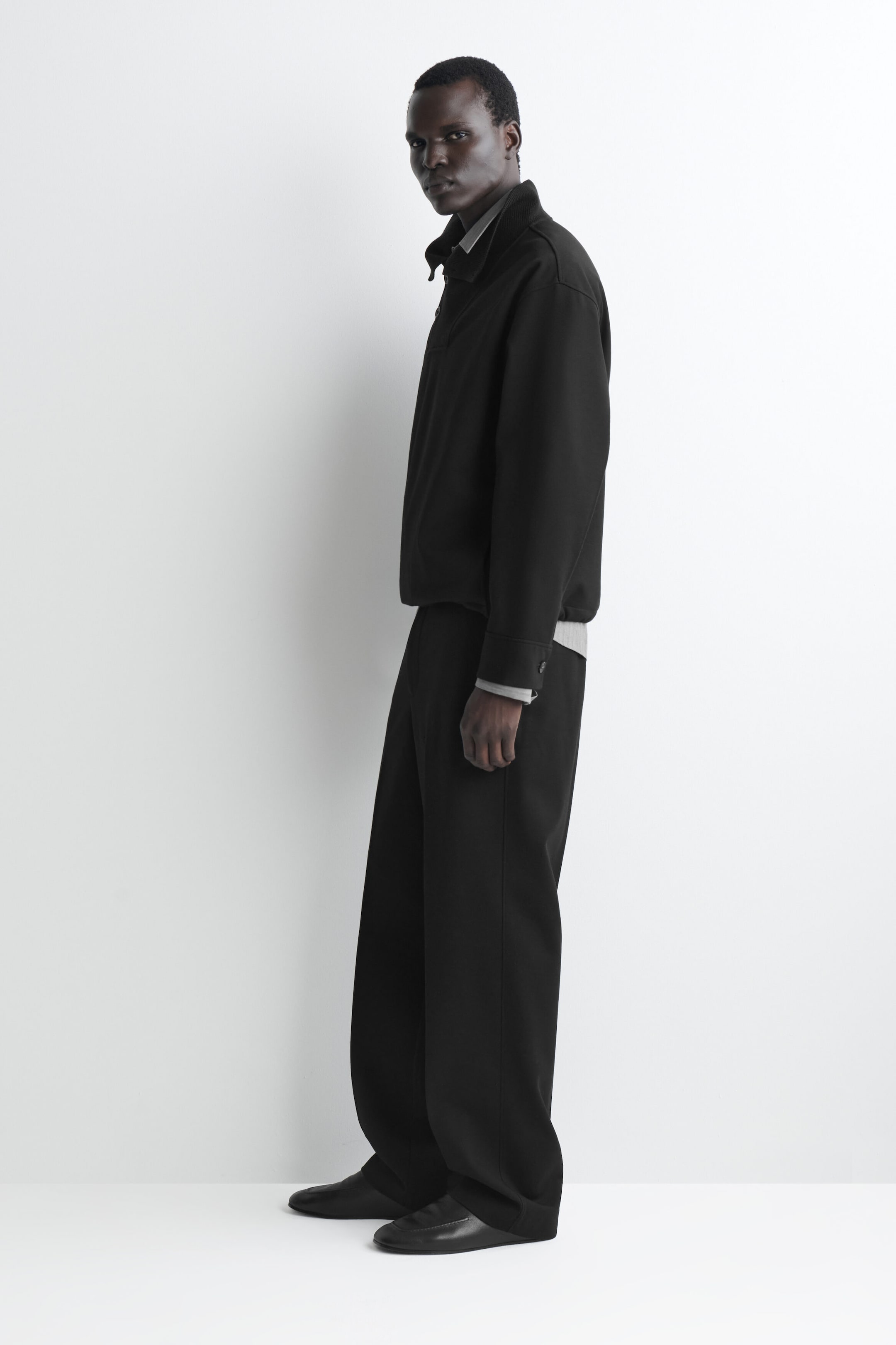 A black jacket and matching wide-leg trousers are shown in a left-facing side profile view. The boxy jacket is layered over a grey shirt, visible at the collar and cuffs, while the relaxed-fit trousers drape over black leather shoes.