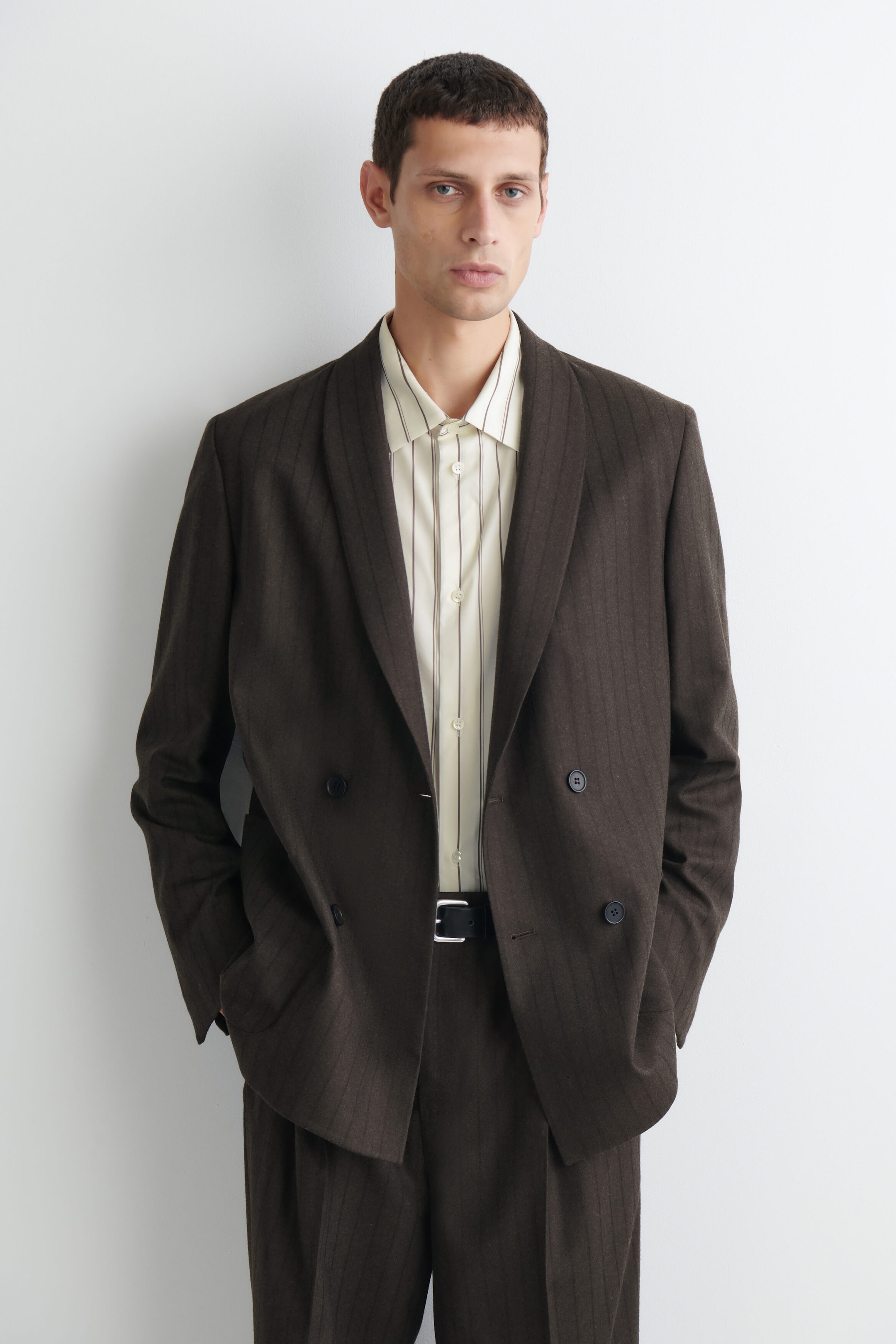 A relaxed-fit, double-breasted brown pinstripe blazer is shown from the front, featuring shawl lapels and a four-button front. The blazer is layered over a light-colored striped shirt and paired with matching trousers. A relaxed-fit, double-breasted brown pinstripe blazer is shown from the front, featuring shawl lapels and a four-button front. The blazer is layered over a light-colored striped shirt and paired with matching trousers.