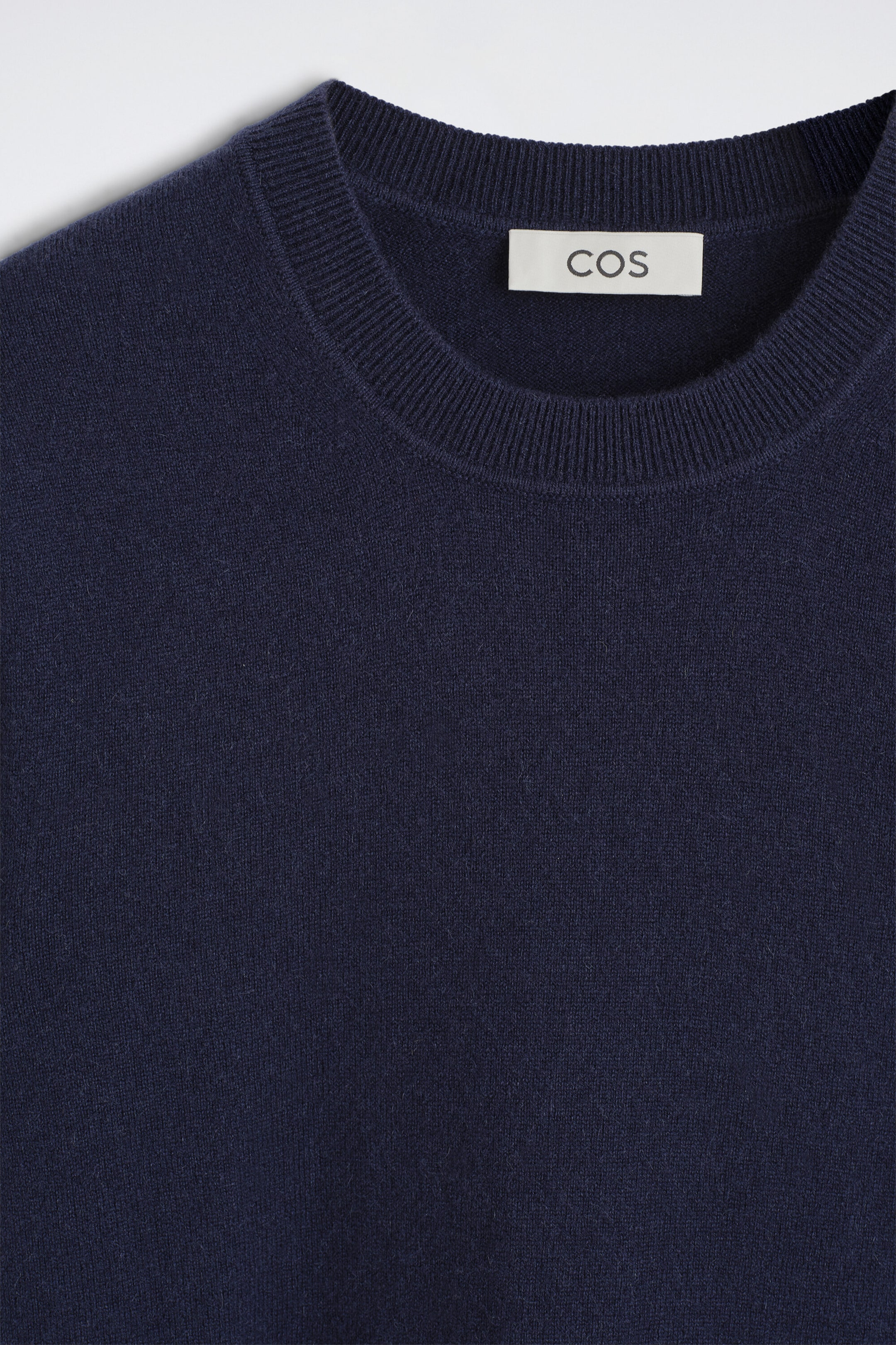A navy blue cashmere crew-neck jumper features a ribbed collar and a fine-knit texture in a close-up front view. A white brand label is visible on the inner neckline. A navy blue cashmere crew-neck jumper features a ribbed collar and a fine-knit texture in a close-up front view. A white brand label is visible on the inner neckline.