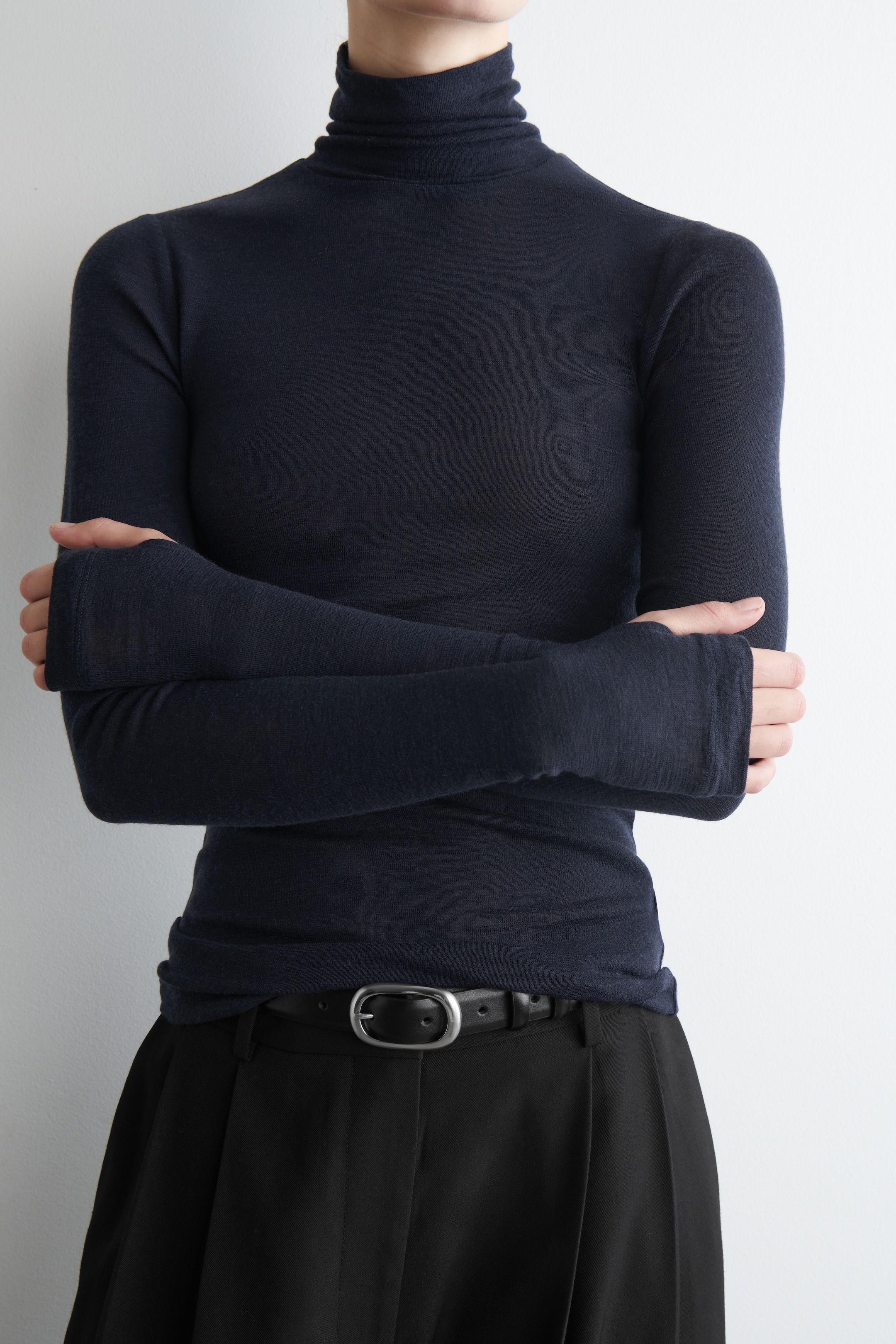 Ingrandisci l'immagine: Un top a collo alto aderente di colore blu scuro è indossato. Questo top in maglia leggera presenta un collo alto arrotolato e maniche lunghe che si estendono oltre le mani. È infilato in pantaloni neri, fissati da una cintura nera con una fibbia color argento.