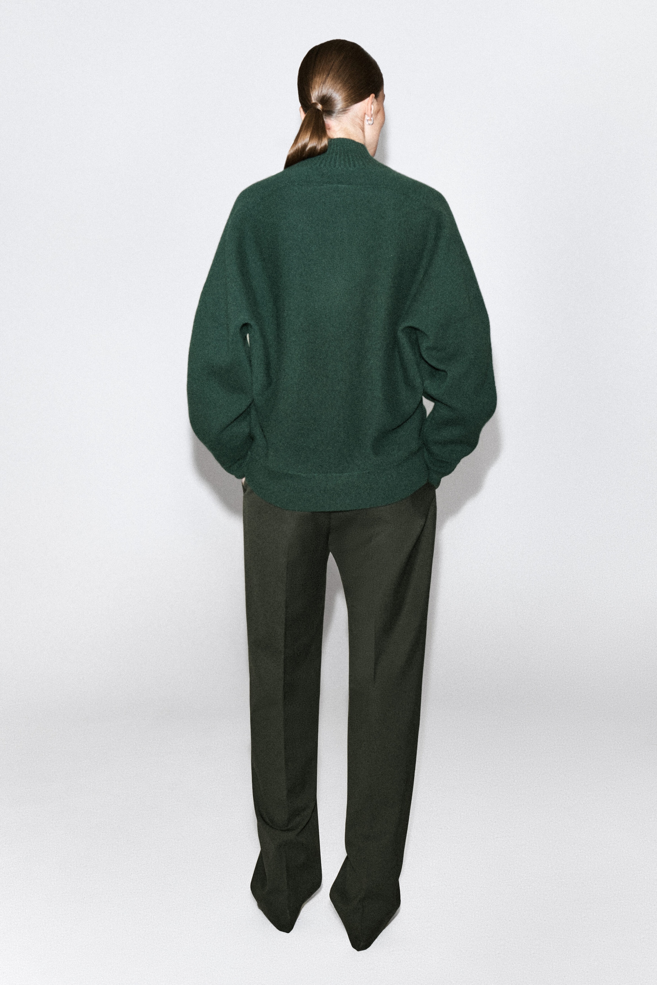 Un chandail en laine bouillie vert foncé est porté par le mannequin, présenté de dos. Le chandail présente une silhouette décontractée et arrondie avec un col cheminée côtelé et un ourlet côtelé, et il est agencé à un pantalon droit foncé. Un chandail en laine bouillie vert foncé est porté par le mannequin, présenté de dos. Le chandail présente une silhouette décontractée et arrondie avec un col cheminée côtelé et un ourlet côtelé, et il est agencé à un pantalon droit foncé.