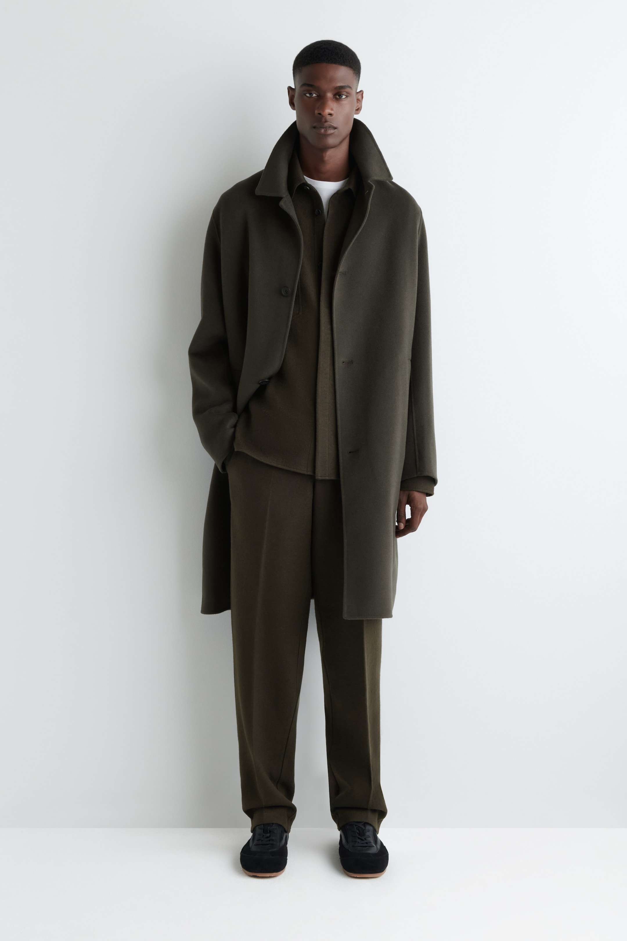Agrandir l'image: Un manteau vert olive foncé est porté ouvert sur une chemise vert olive assortie et un pantalon large, formant un ensemble monochromatique. Le manteau présente un large col montant et une silhouette longue et ample, et des baskets noires avec des semelles marron clair visibles complètent le look.