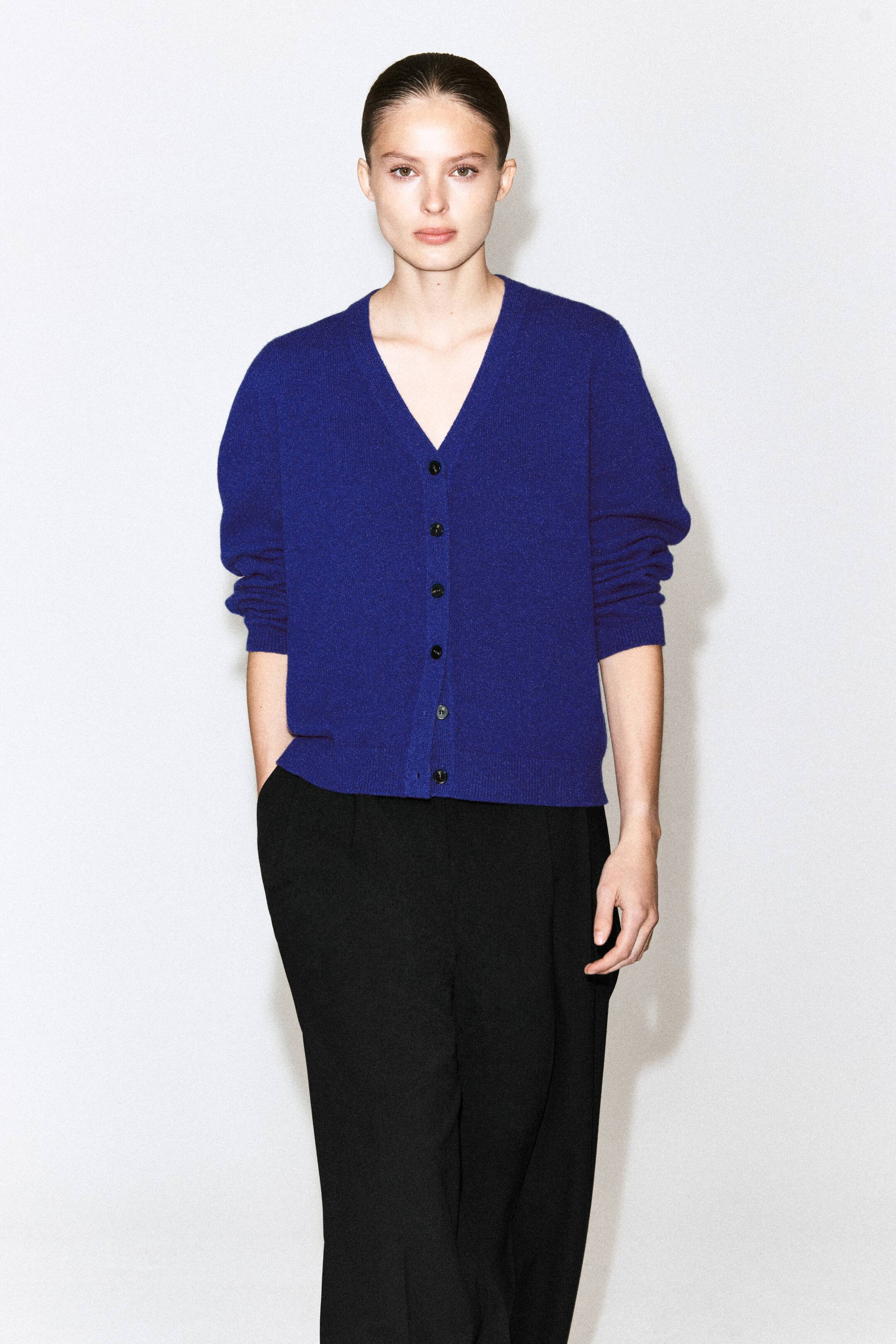 View larger image: This royal blue merino wool V-neck cardigan is worn by the model and features a front button fastening and ribbed trims at the cuffs and hem. The buttoned cardigan is paired with black trousers, with the model resting one hand in a pocket.