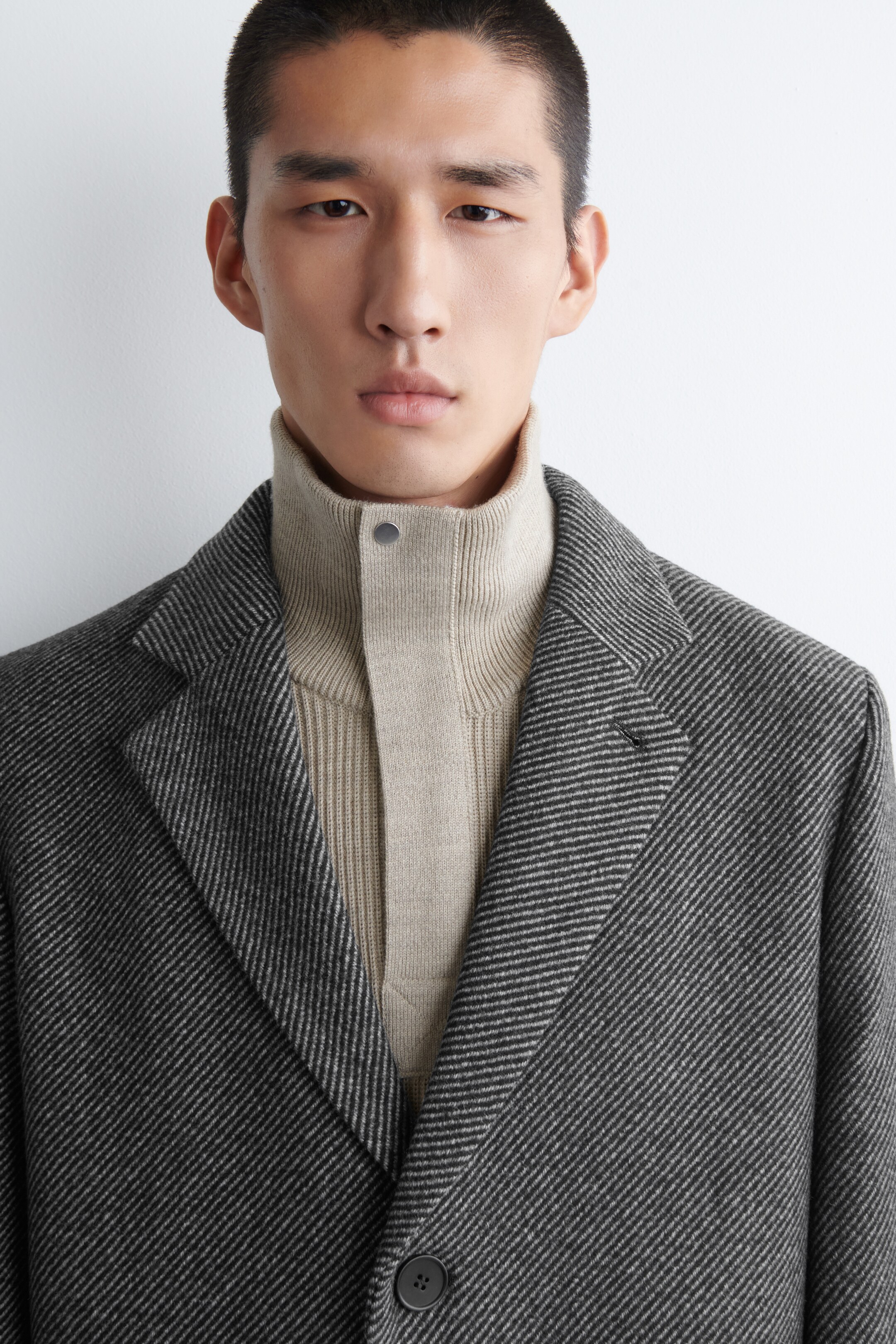 Agrandir l'image: Un manteau gris foncé est porté sur un col roulé beige, légèrement tourné vers l'avant. Le manteau présente de larges revers à cran et un tissage en diagonales. Le col roulé côtelé affiche un panneau avant lisse avec une seule fermeture à bouton-pression.