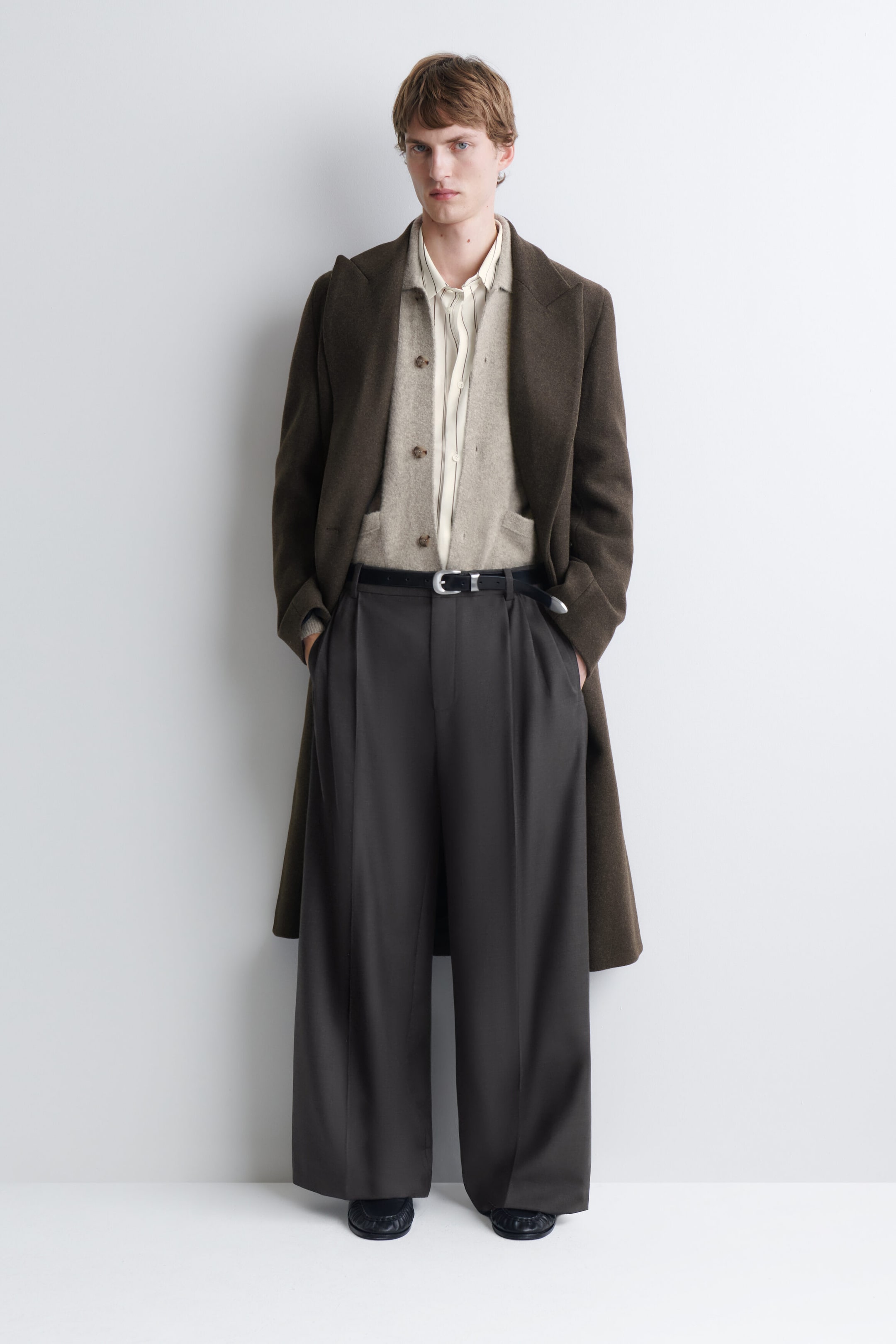 A beige brushed-knit cardigan is being worn by the model, featuring a classic collar, front button closure, and patch pockets. The cardigan is layered over a striped button-down shirt and under a long, dark brown overcoat, styled with dark wide-leg trousers. A beige brushed-knit cardigan is being worn by the model, featuring a classic collar, front button closure, and patch pockets. The cardigan is layered over a striped button-down shirt and under a long, dark brown overcoat, styled with dark wide-leg trousers.