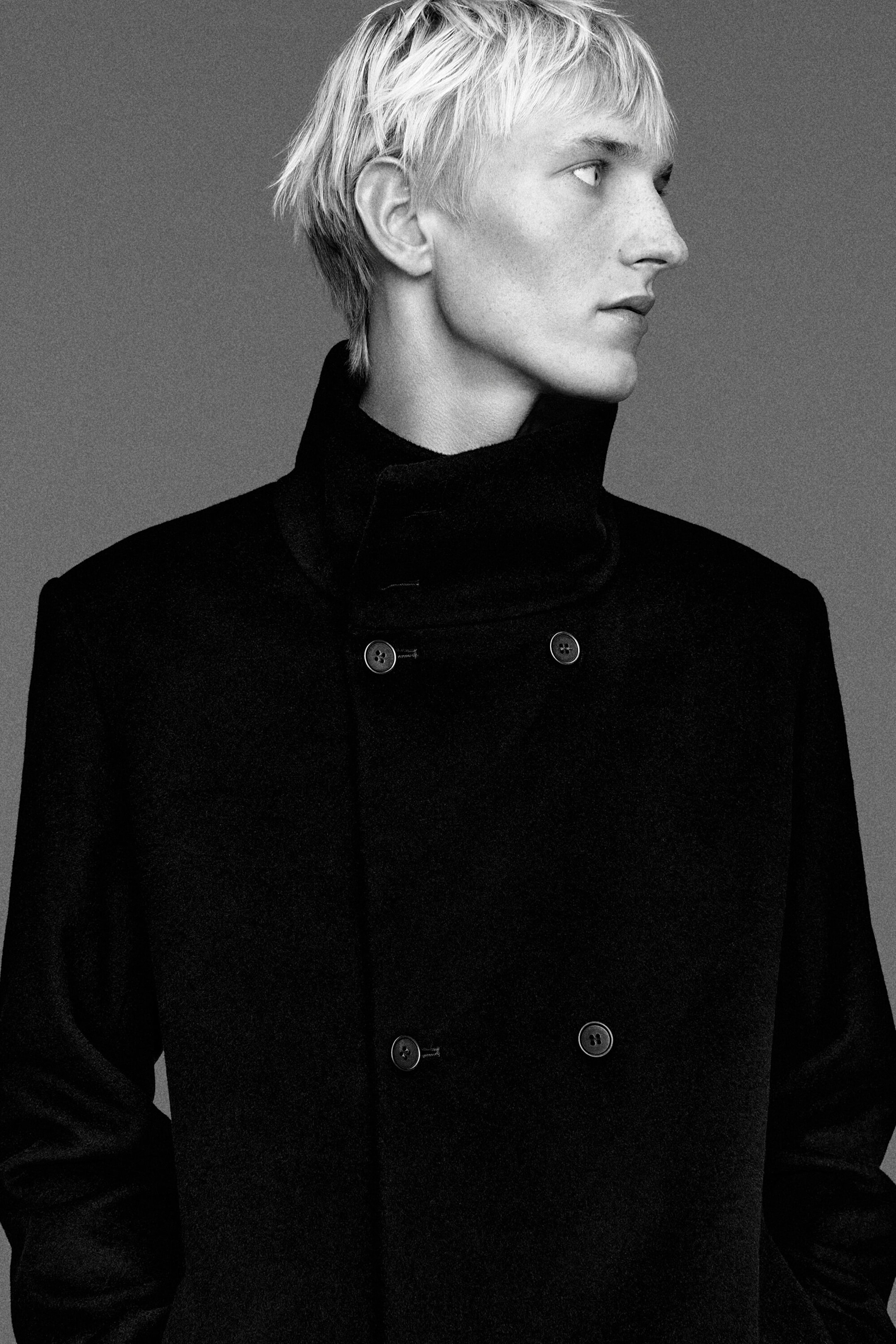 Un mannequin porte un manteau foncé croisé, vu de face, mettant en valeur son col cheminée haut, ses épaules structurées et sa fermeture boutonnée soignée. Ce gros plan souligne les lignes épurées et la texture tailleur du vêtement. Un mannequin porte un manteau foncé croisé, vu de face, mettant en valeur son col cheminée haut, ses épaules structurées et sa fermeture boutonnée soignée. Ce gros plan souligne les lignes épurées et la texture tailleur du vêtement.