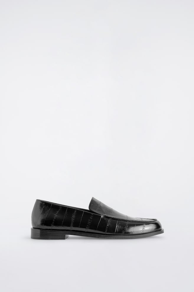 Un mocassin noir en cuir est présenté de profil, mettant en valeur une finition polie avec un motif embossé effet peau d’anguille. La chaussure est conçue avec un bout arrondi, un petit talon empilé et une semelle plate. - 1