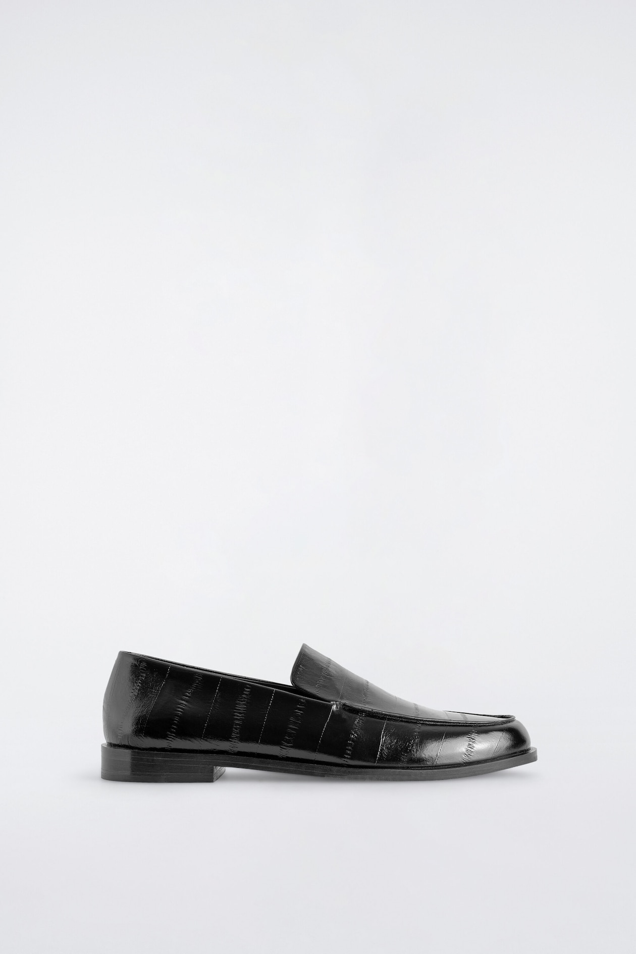 A single black leather loafer is shown in side profile, showcasing a polished finish with an embossed eel-effect motif. The shoe is designed with a rounded toe, a low stacked heel, and a flat sole.