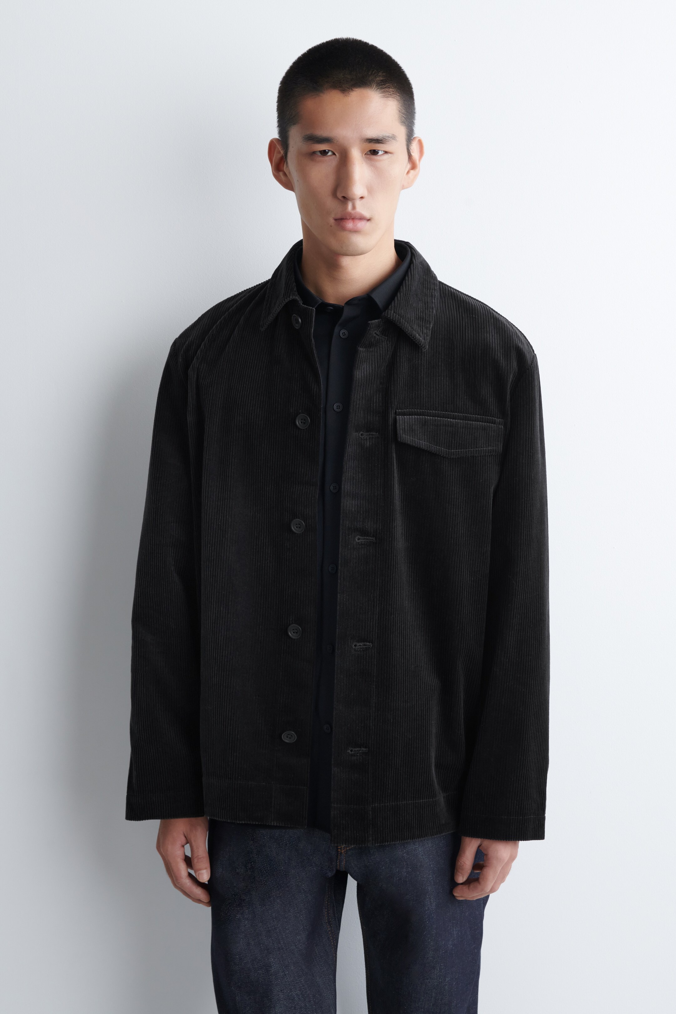 View larger image: A dark corduroy jacket is facing forward, featuring a pointed collar, a button-front closure with dark buttons, long sleeves, and a single patch pocket on the left chest. The jacket is being worn over a dark collared shirt.