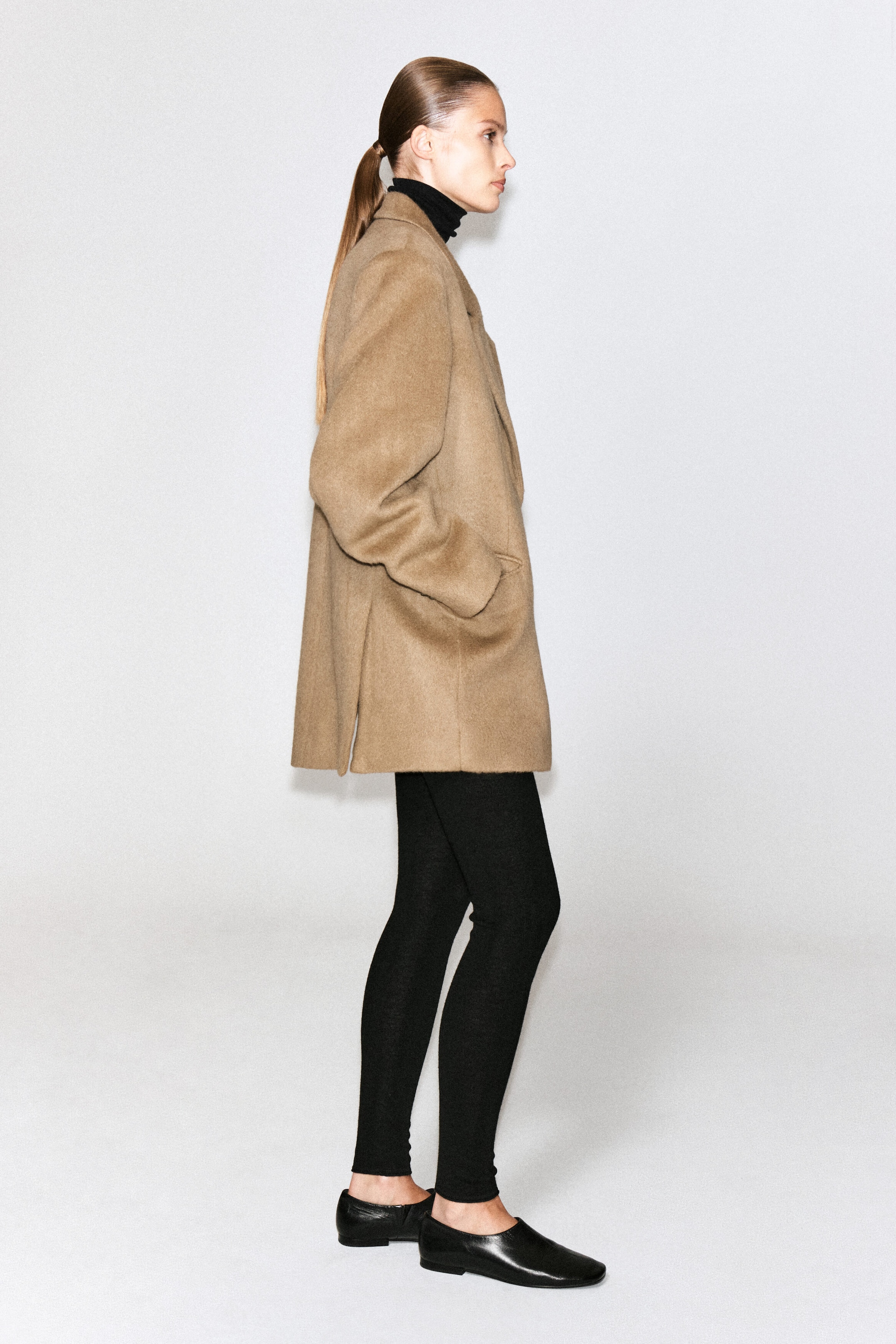 A brushed wool blazer in a camel-brown shade is worn by the model, who stands in profile with her hands in the side pockets. The mid-length, structured blazer features a side vent at the hem and is styled over a black turtleneck and black leggings. A brushed wool blazer in a camel-brown shade is worn by the model, who stands in profile with her hands in the side pockets. The mid-length, structured blazer features a side vent at the hem and is styled over a black turtleneck and black leggings.