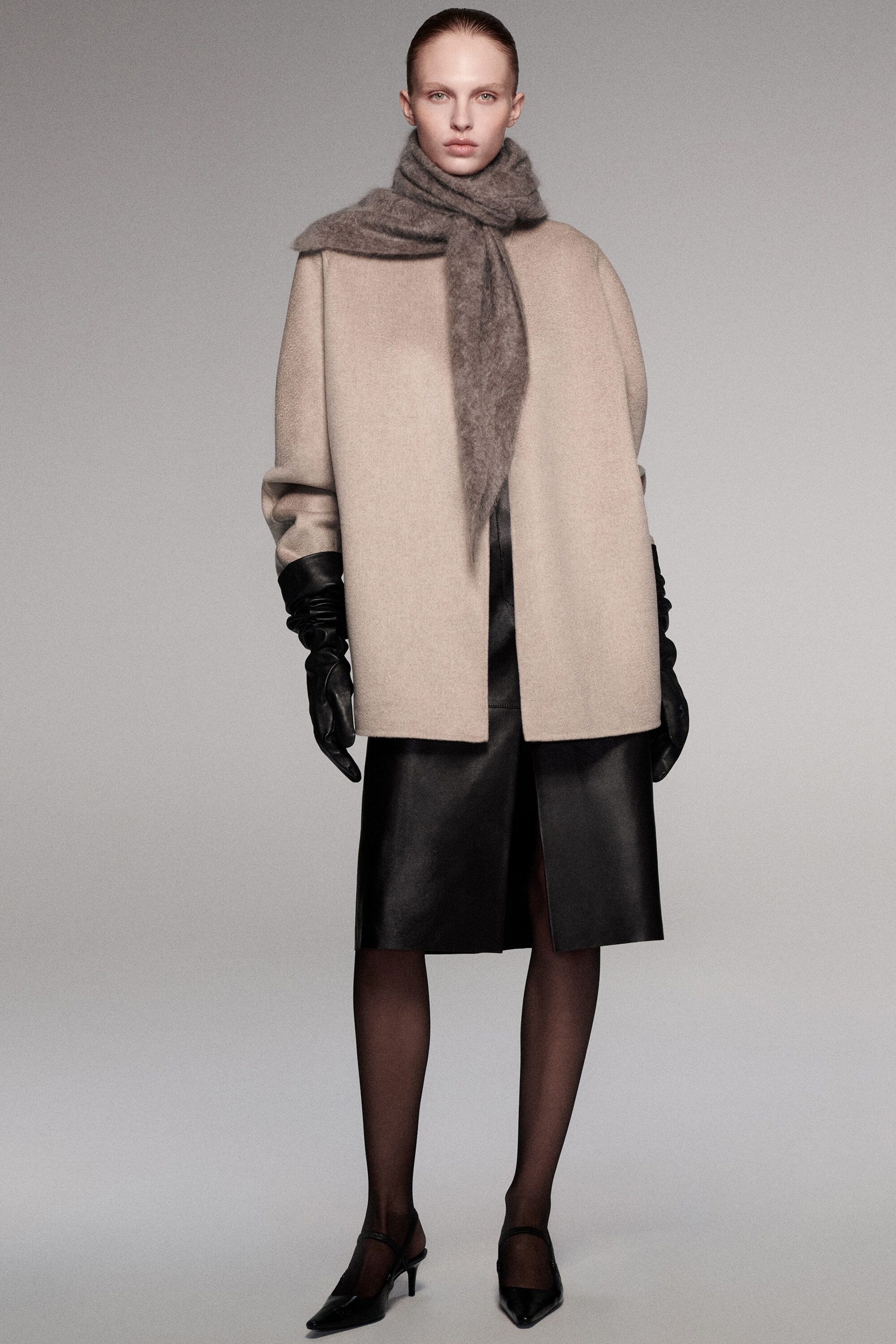 This double-faced cashmere funnel-neck jacket in light beige is styled in a relaxed, boxy fit from the front, featuring an open-front design and wide sleeves. It is paired with a gray textured scarf wrapped around the neck, long black leather gloves tucked into the sleeves, and a black leather skirt visible beneath the straight hem. This double-faced cashmere funnel-neck jacket in light beige is styled in a relaxed, boxy fit from the front, featuring an open-front design and wide sleeves. It is paired with a gray textured scarf wrapped around the neck, long black leather gloves tucked into the sleeves, and a black leather skirt visible beneath the straight hem.