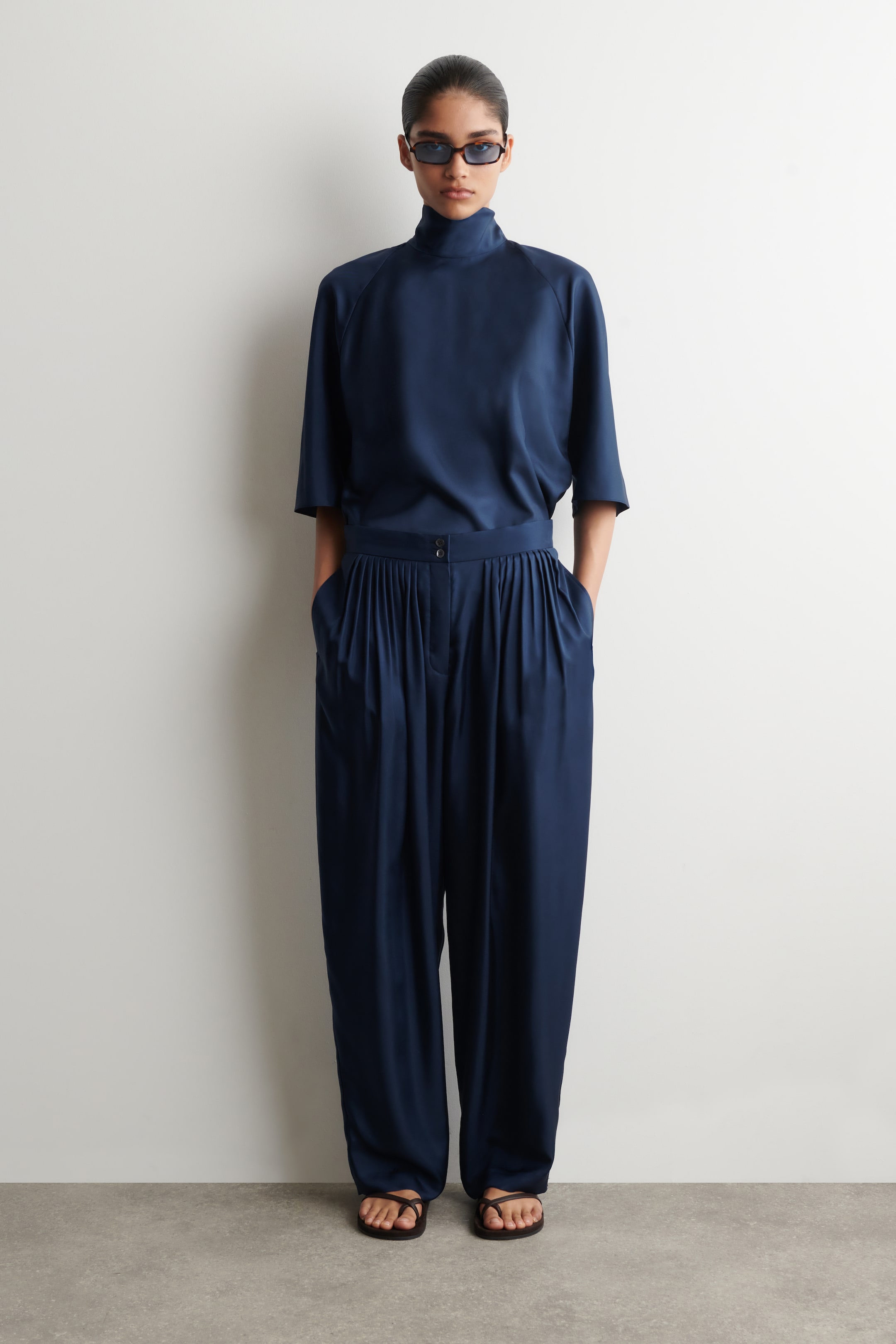 Agrandir l'image: Un ensemble assorti bleu foncé se compose d'un haut à col montant avec des manches raglan longueur coude, rentré dans un pantalon taille haute à jambes larges. Le pantalon présente des plis profonds à l'avant et une fermeture à deux boutons, le tout confectionné dans un tissu fluide et brillant.