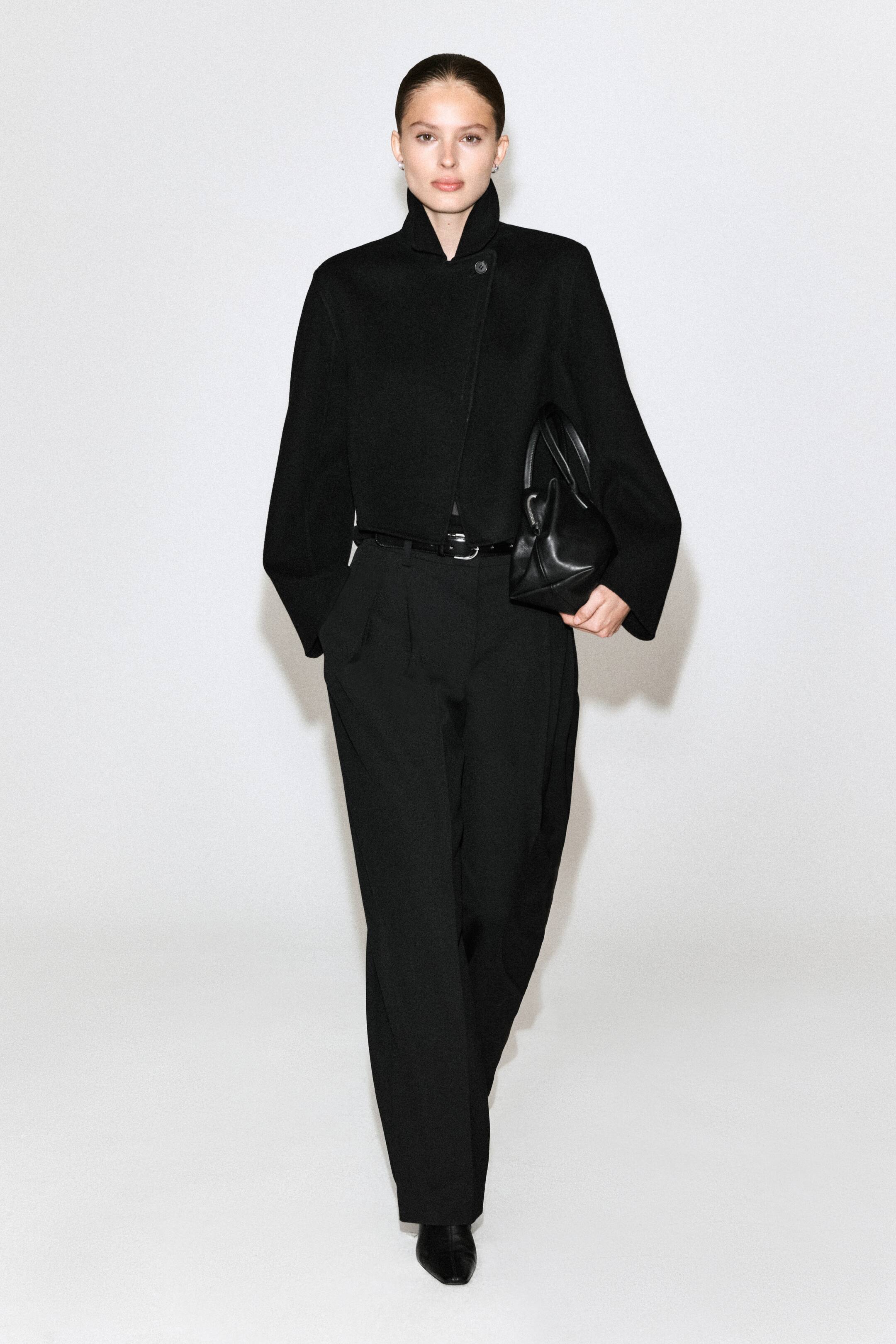 Agrandir l'image: Un mannequin porte une veste courte noire dotée d’un col montant et d’une fermeture asymétrique à boutons, associée à un pantalon noir plissé à taille haute et à jambe large, maintenu par une fine ceinture noire. Elle complète le look avec des chaussures noires à bout pointu et tient un petit sac à main structuré noir.