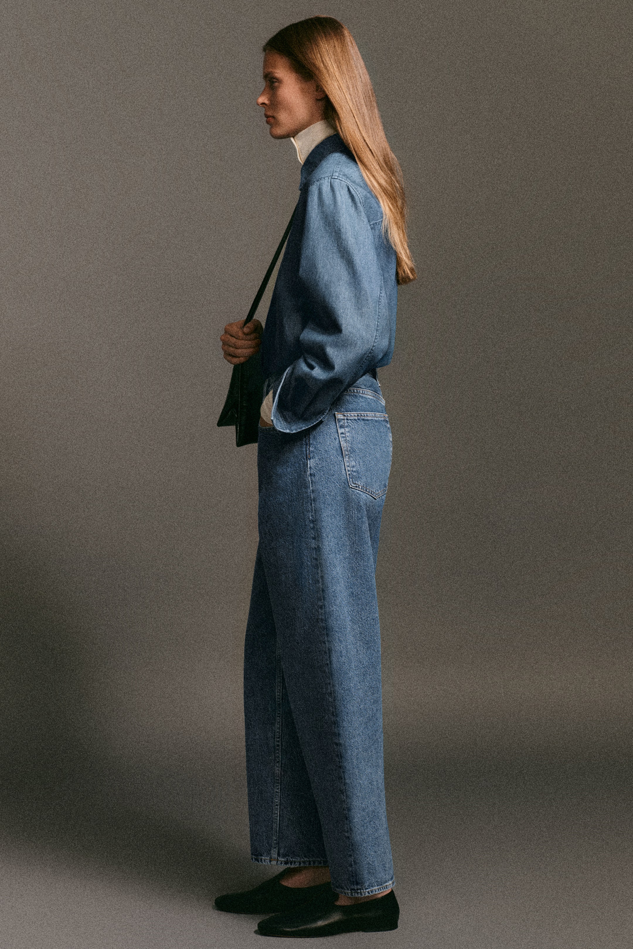 Ver imagen más grande: La camisa vaquera de corte largo y los jeans bay de pierna barrel aparecen en vista de perfil como un conjunto coordinado de lavado medio. La camisa presenta mangas largas sobrepuestas a un top crema de cuello alto, mientras que los jeans de tiro alto muestran una silueta curva de pierna barrel y un bolsillo trasero clásico.