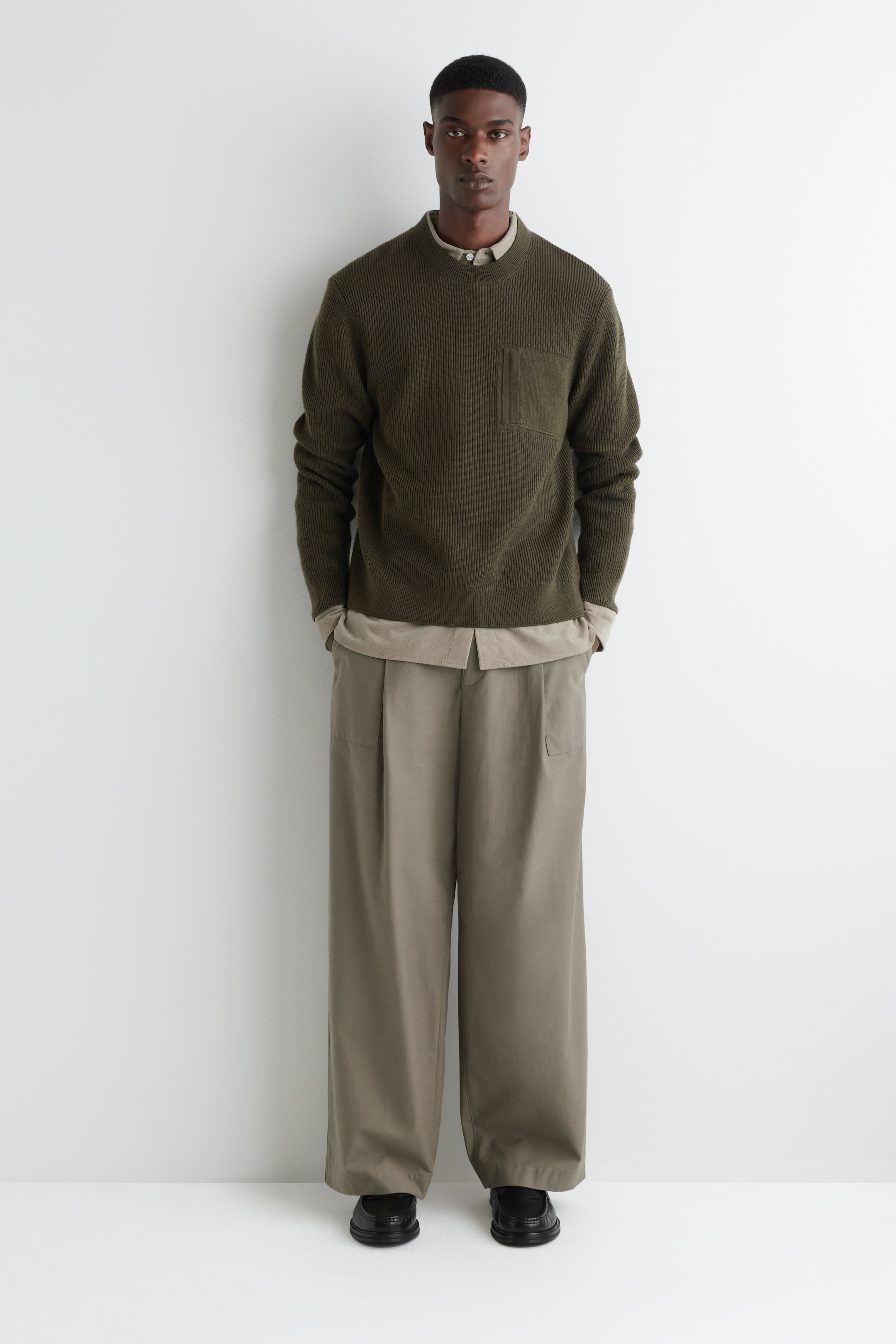 Visualizza immagine più grande: Un maglione verde oliva scuro con scollo a girocollo, rivolto in avanti, presenta una trama a maglia a coste verticali e una tasca quadrata sul petto sinistro. È stratificato su una camicia con colletto color beige chiaro, visibile al collo e all'orlo, ed è abbinato a pantaloni a gamba larga color cachi chiaro.