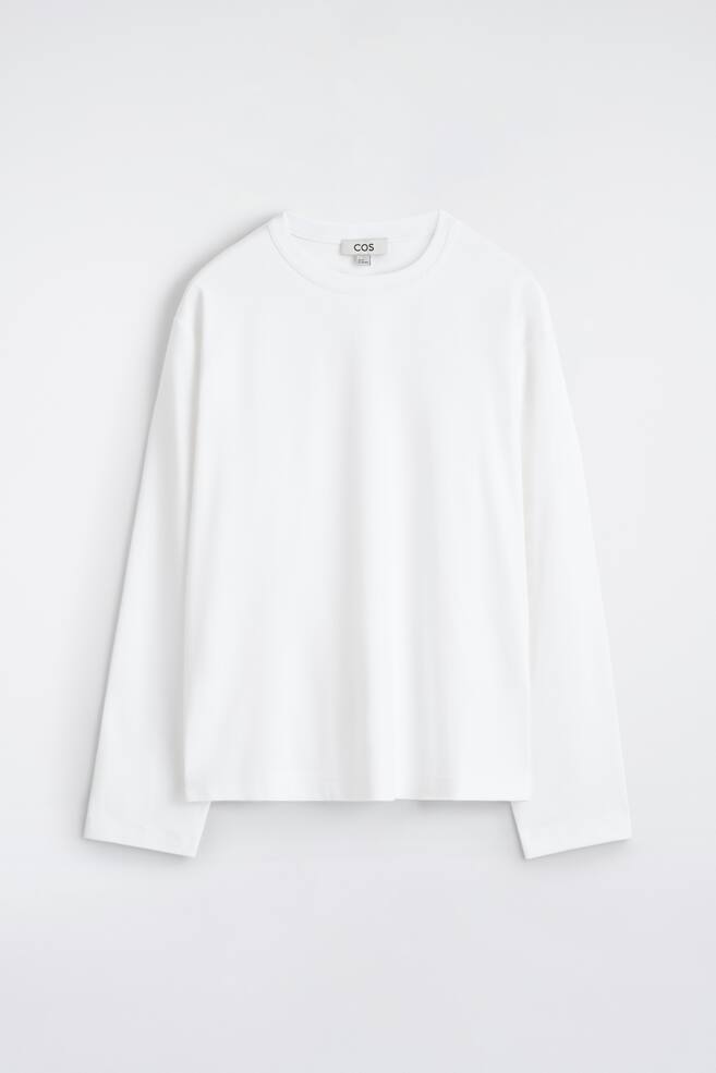 A white long-sleeved T-shirt is shown from the FRONT, featuring a relaxed fit and a classic crew neck. A small 'COS' brand tag is visible at the back of the neck. - 1