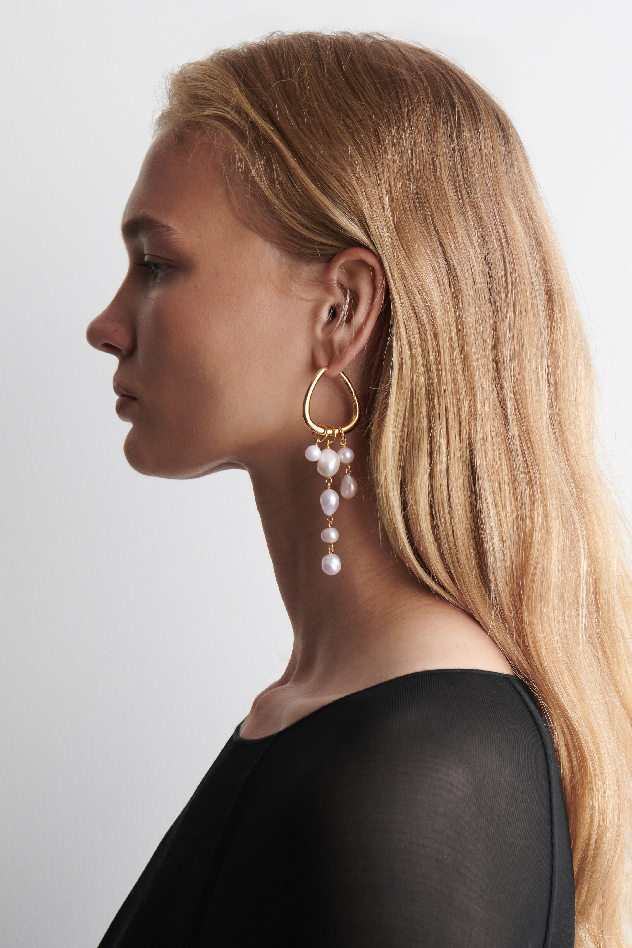 View larger image: A gold-toned earring is dangling from an ear, facing left in profile view. It is featuring an irregular loop at the top, from which multiple strands of varying-sized baroque pearls are hanging.
