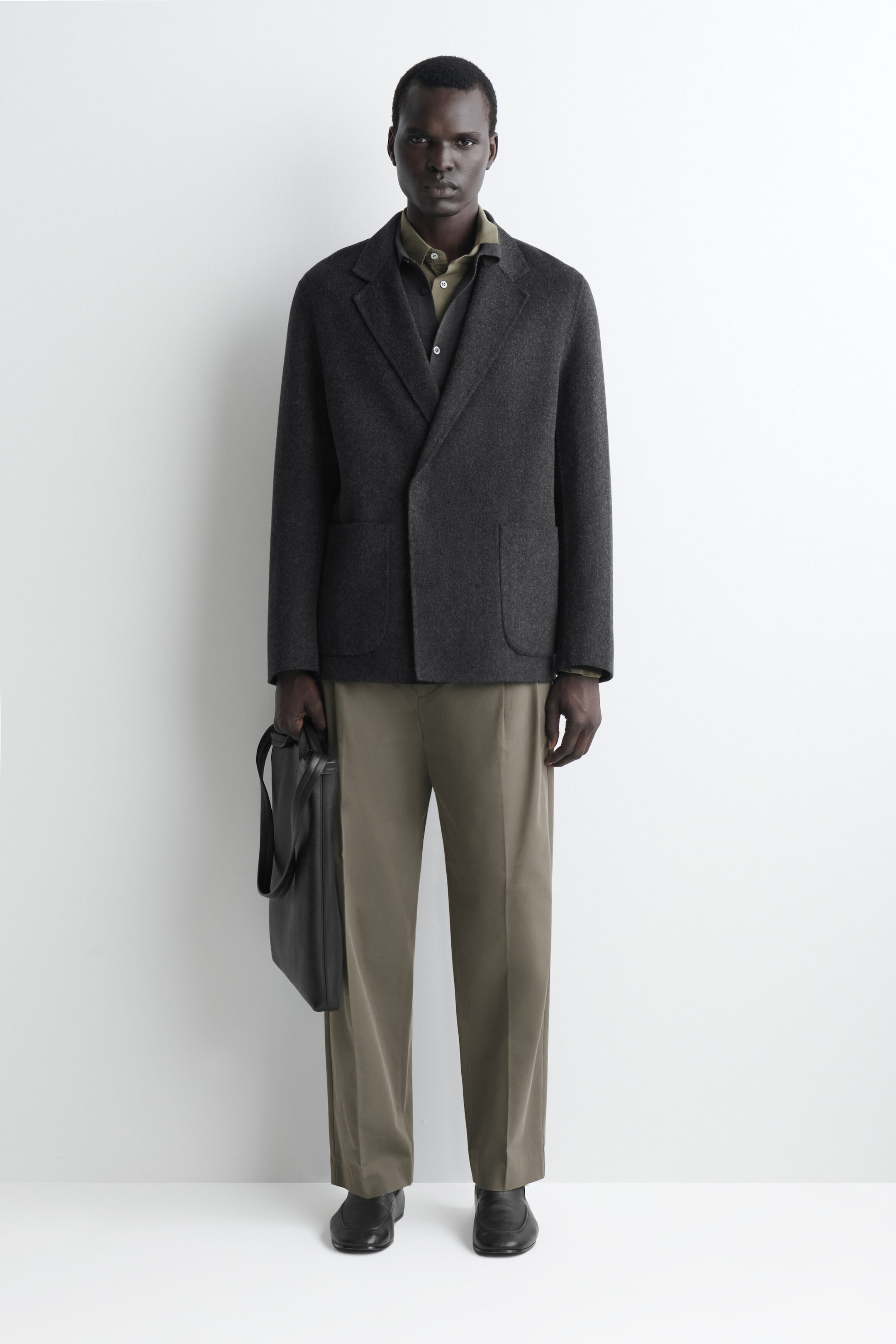 This dark grey, unstructured double-breasted blazer is shown from the front, highlighting its relaxed fit, notch lapels and clean placket. The blazer features two front patch pockets and is styled over an olive-green shirt with matching trousers. This dark grey, unstructured double-breasted blazer is shown from the front, highlighting its relaxed fit, notch lapels and clean placket. The blazer features two front patch pockets and is styled over an olive-green shirt with matching trousers.