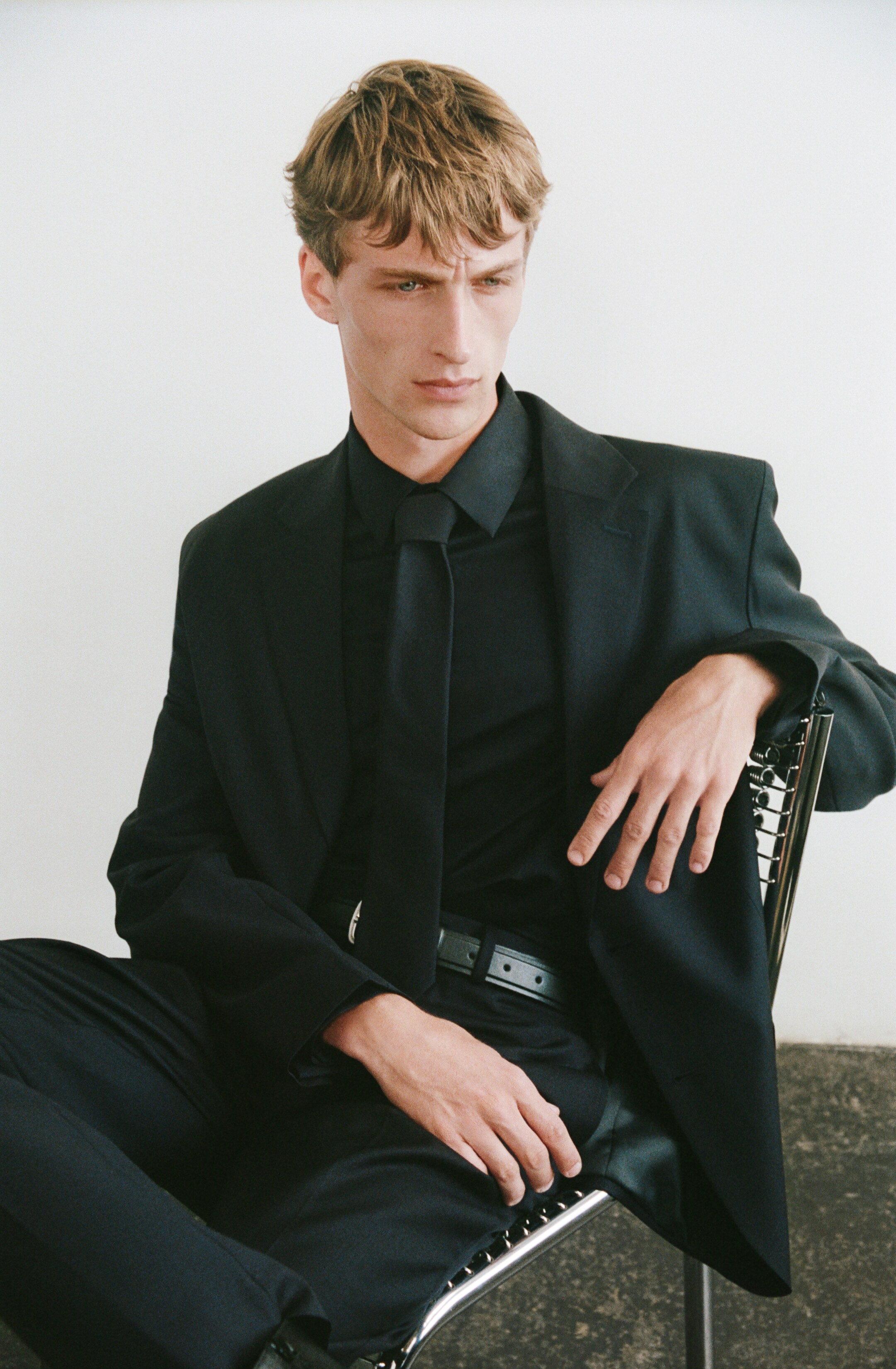 Größeres Bild anzeigen: Ein schwarzer einreihiger Blazer mit Kerbrevers wird über einem schwarzen langärmeligen Hemd mit spitzem Kragen und einer schwarzen Krawatte getragen. Passende schwarze Hosen werden ebenfalls getragen, gesichert durch einen schwarzen Gürtel mit einer silbernen Schnalle.