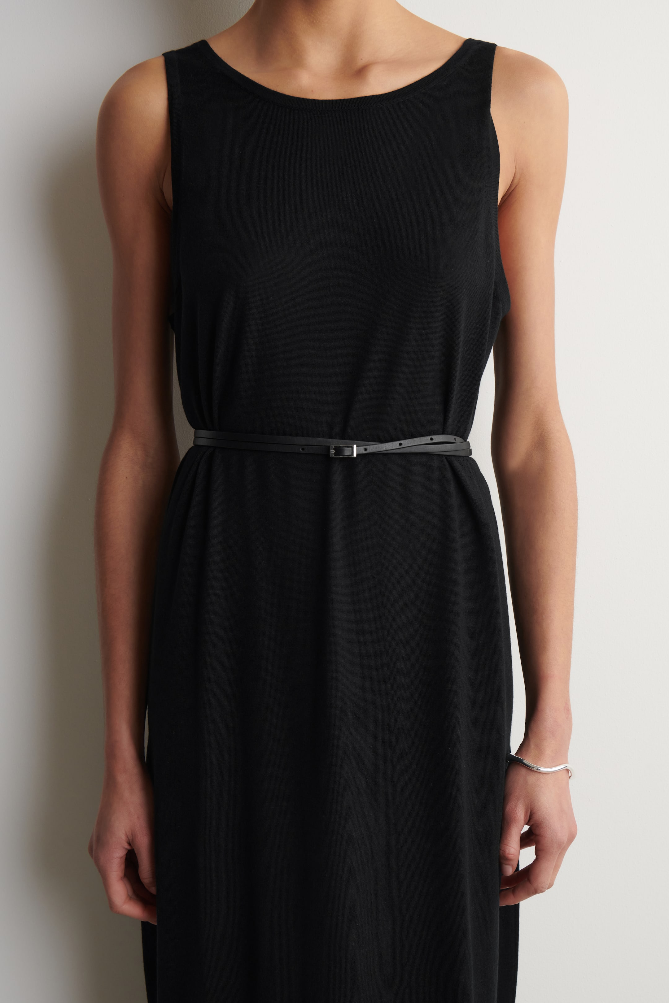 Größeres Bild ansehen: Ein schwarzes ärmelloses Kleid mit rundem Ausschnitt und weichem, fließendem Stoff. Ein dünner schwarzer Gürtel mit einer kleinen silbernen Schnalle wird zweimal um die Taille gewickelt und verleiht dem Kleid eine taillierte Silhouette.