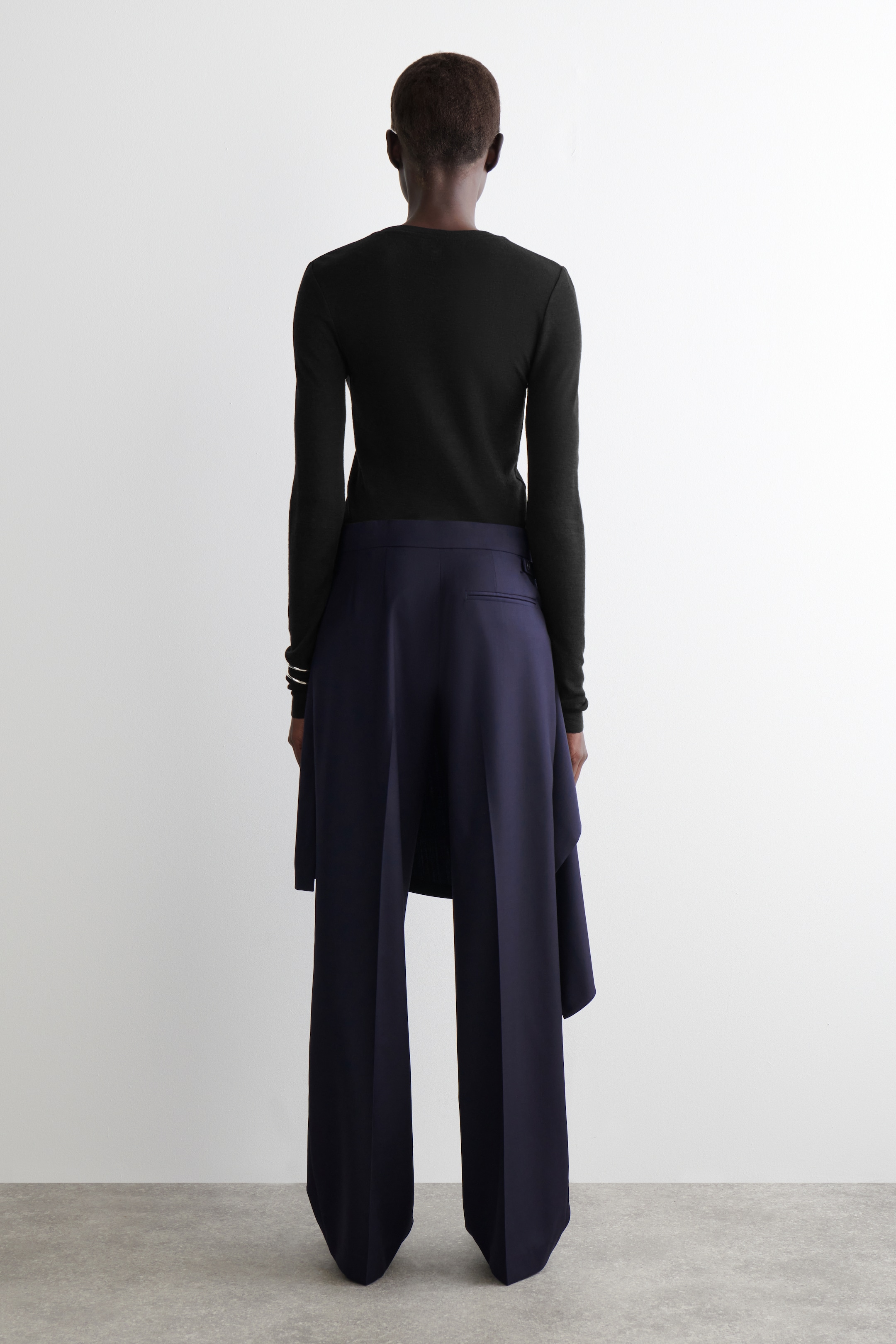 View larger image: A black long-sleeved top is being worn, viewed from the back. It is paired with navy wide-leg trousers that are featuring a distinctive draped overlay, creating a fluid, layered effect over the legs.