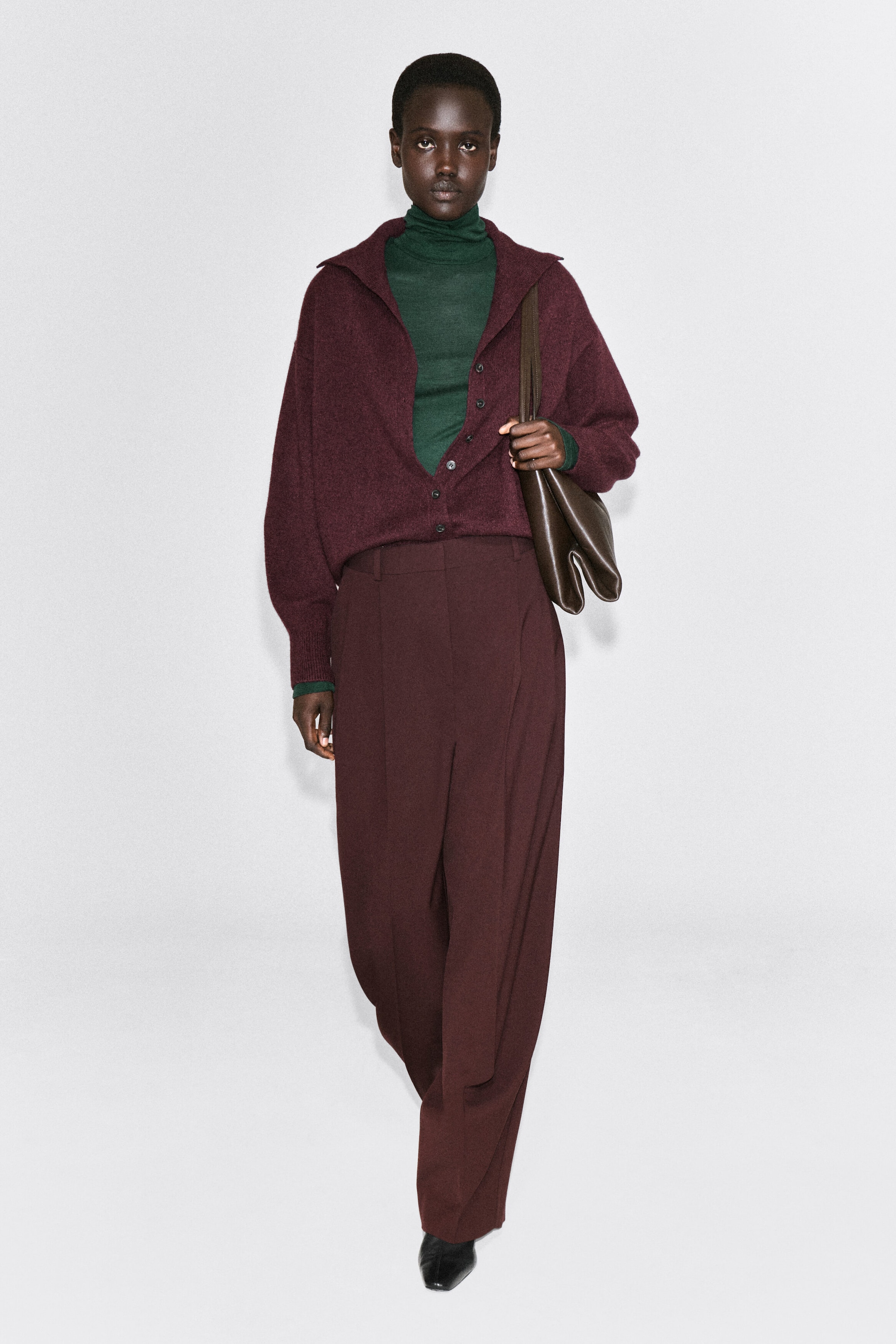 The burgundy cashmere cardigan, facing front, is styled partially unbuttoned to form a wide collar, showcasing its relaxed fit, sculpted sleeves, and long ribbed cuffs. It is layered over a dark green roll-neck top and paired with matching burgundy wide-leg trousers. The burgundy cashmere cardigan, facing front, is styled partially unbuttoned to form a wide collar, showcasing its relaxed fit, sculpted sleeves, and long ribbed cuffs. It is layered over a dark green roll-neck top and paired with matching burgundy wide-leg trousers.