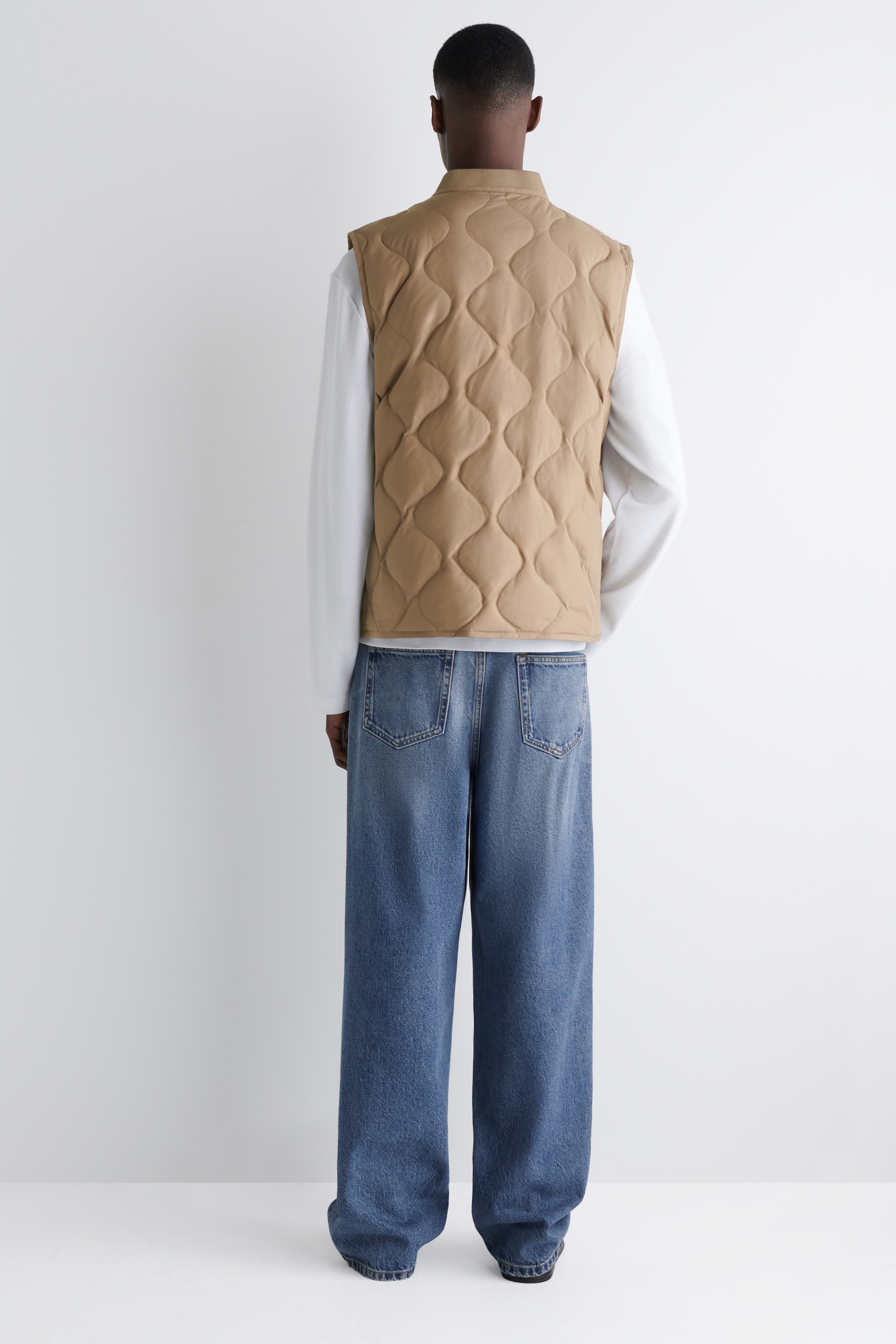 Agrandir l'image: Un gilet matelassé beige est présenté de dos, avec un motif matelassé ondulé distinctif et un col montant. Il est superposé à une chemise blanche à manches longues.