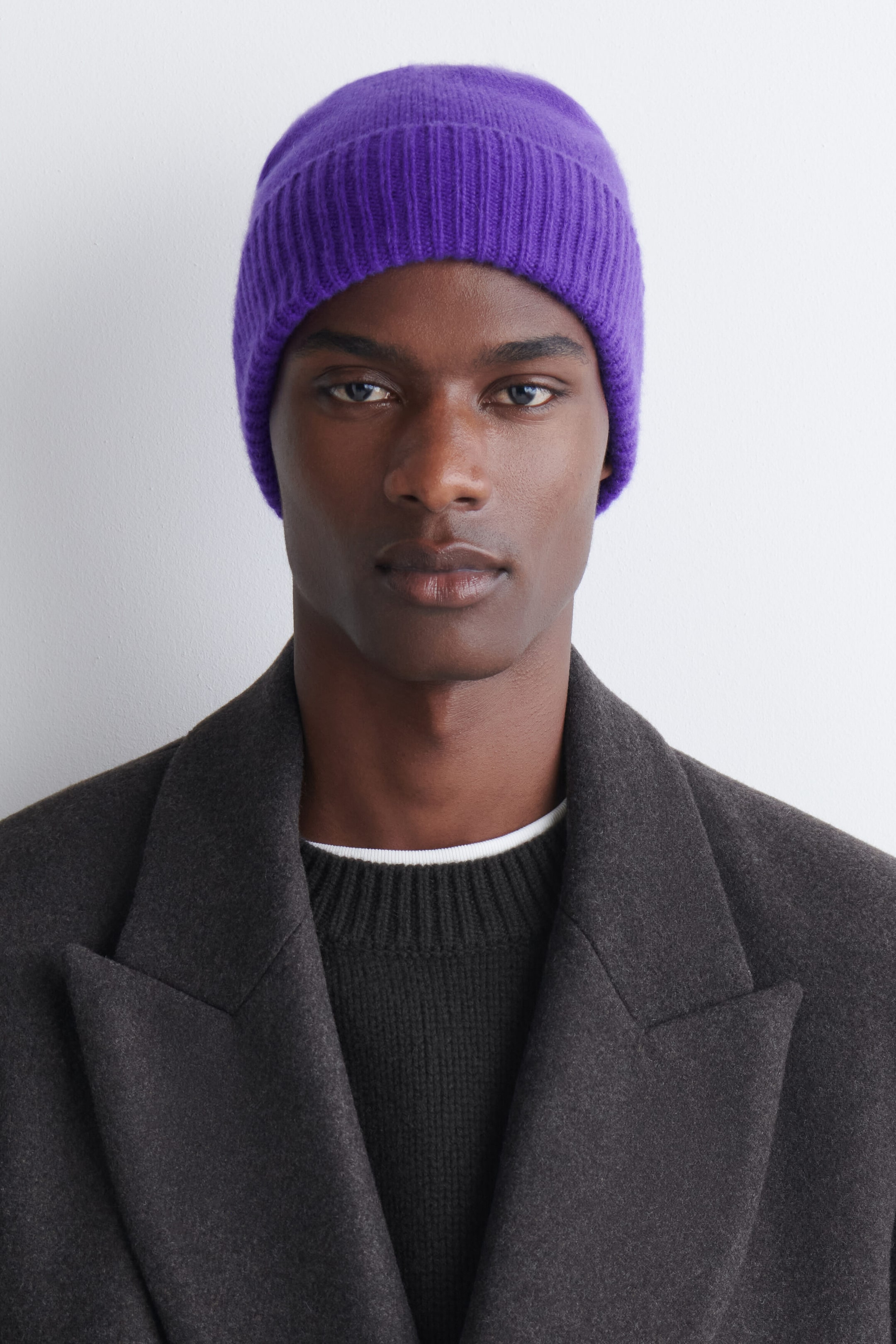 Eine Beanie aus reinem Kaschmir in lebhaftem Violett wird von vorne getragen und zeichnet sich durch eine gerippte Strickstruktur sowie einen breiten umgeschlagenen Bund aus. Die eng anliegende Beanie sitzt tief auf der Stirn und ist zusammen mit einem dunkelgrauen Mantel und einem schwarzen Rundhals-Pullover gestylt. Eine Beanie aus reinem Kaschmir in lebhaftem Violett wird von vorne getragen und zeichnet sich durch eine gerippte Strickstruktur sowie einen breiten umgeschlagenen Bund aus. Die eng anliegende Beanie sitzt tief auf der Stirn und ist zusammen mit einem dunkelgrauen Mantel und einem schwarzen Rundhals-Pullover gestylt.