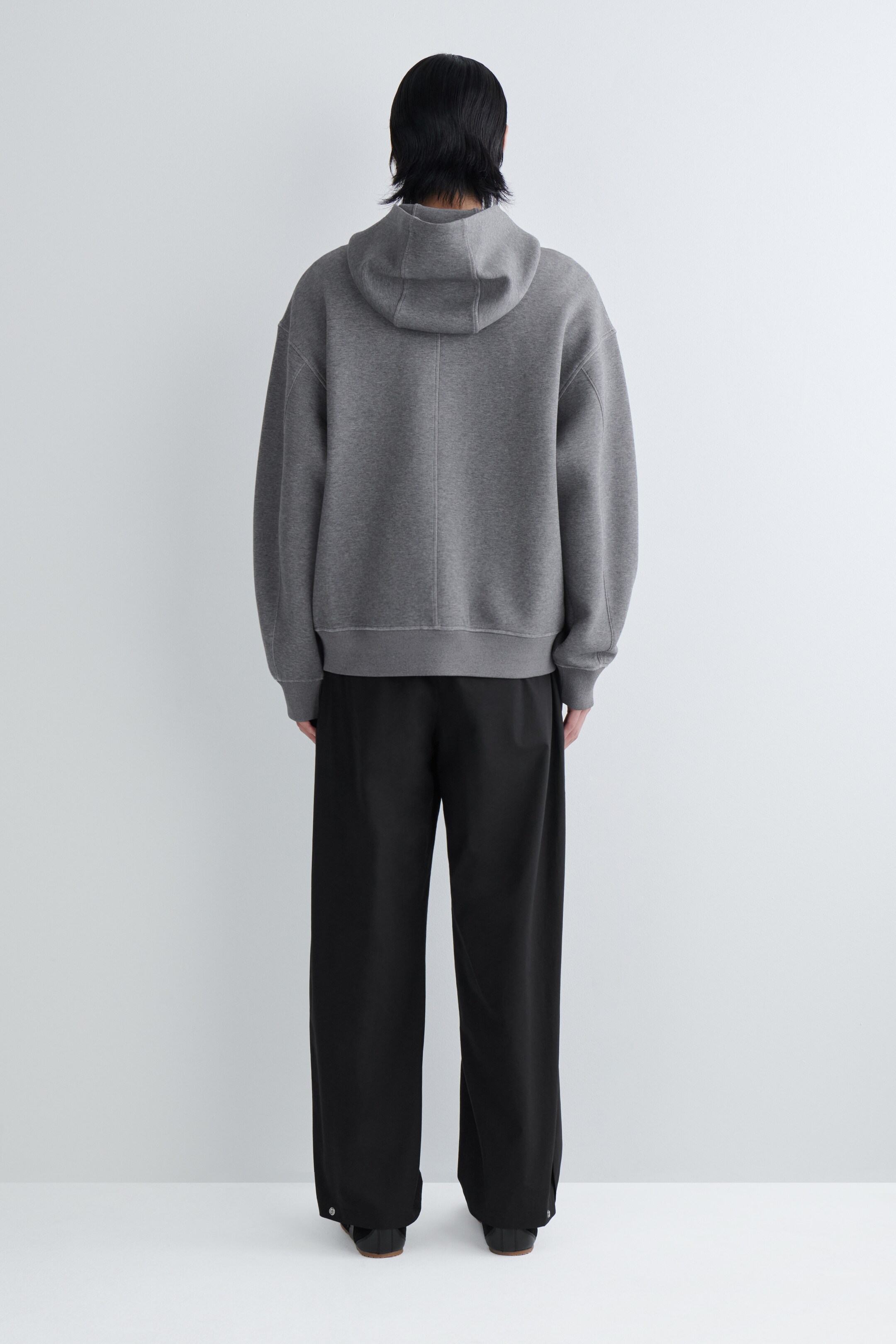 Zobrazit větší obrázek: Šedá oversized mikina s kapucí je prezentována zezadu, s viditelnou kapucí, žebrovanými manžetami, žebrovaným lemem a středovým švem na zádech. Černé kalhoty s širokými nohavicemi jsou nošeny s mikinou, mají uvolněný střih a detail knoflíku u každého kotníku.