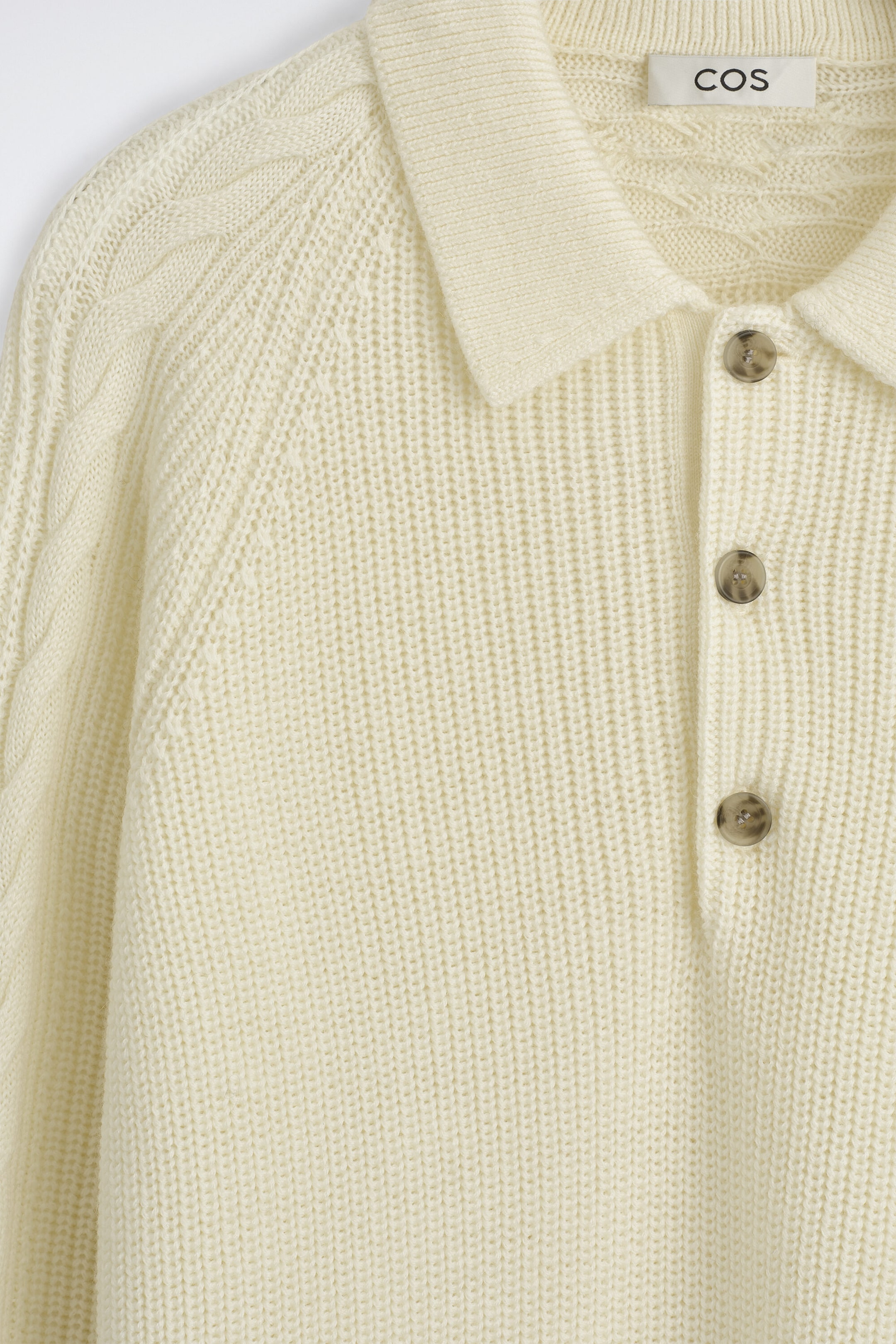 A cream-colored chunky-knit polo sweater is shown in a close-up front view, featuring a ribbed texture, a classic collar, and a three-button placket. The sweater showcases intricate cable-knit detailing along the raglan shoulder seams. A cream-colored chunky-knit polo sweater is shown in a close-up front view, featuring a ribbed texture, a classic collar, and a three-button placket. The sweater showcases intricate cable-knit detailing along the raglan shoulder seams.