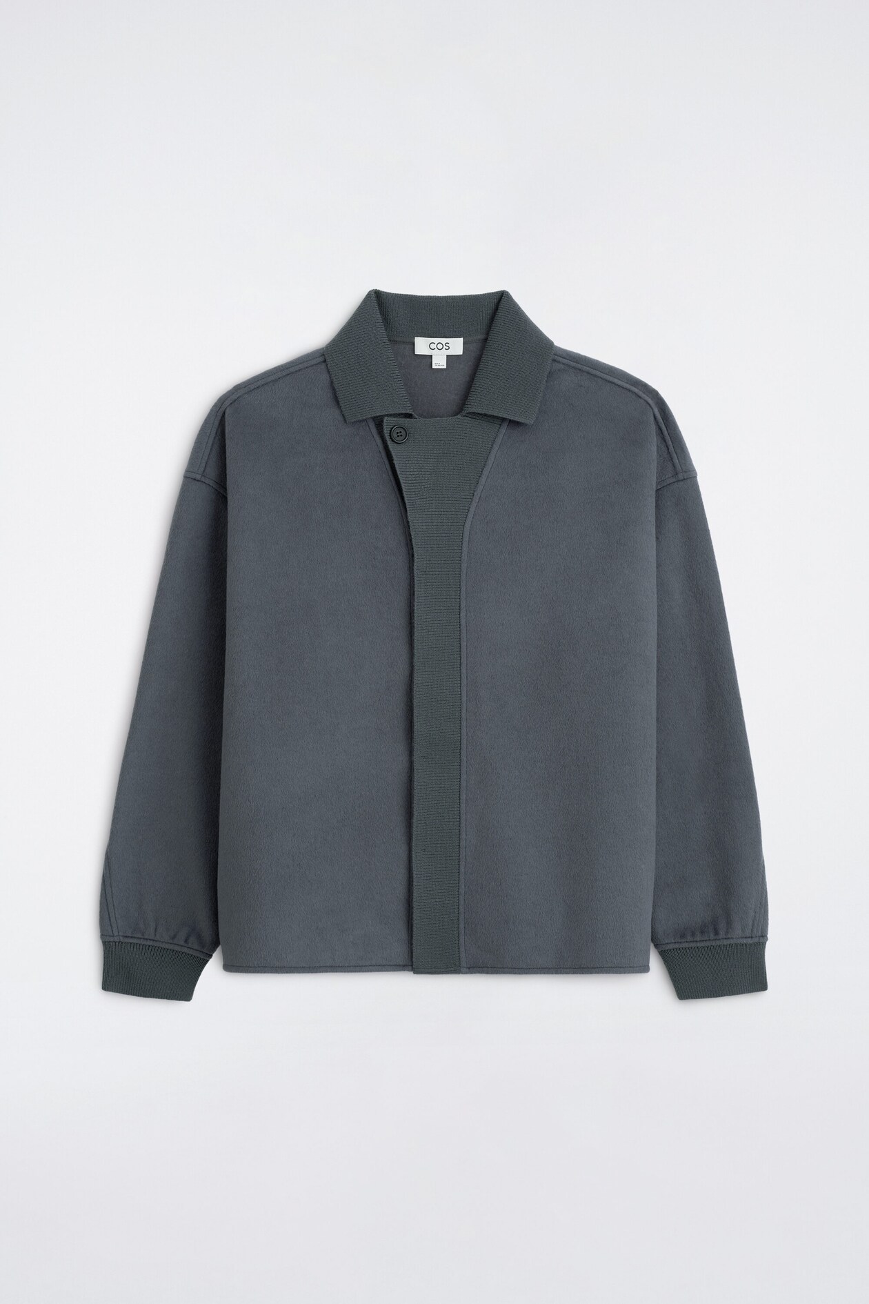 Une veste gris foncé est présentée de face, affichant une texture feutrée avec un col à revers côtelé et des poignets côtelés. Elle présente une fermeture asymétrique à un seul bouton à l’encolure ainsi qu’une patte de boutonnage centrale large, côtelée verticalement.