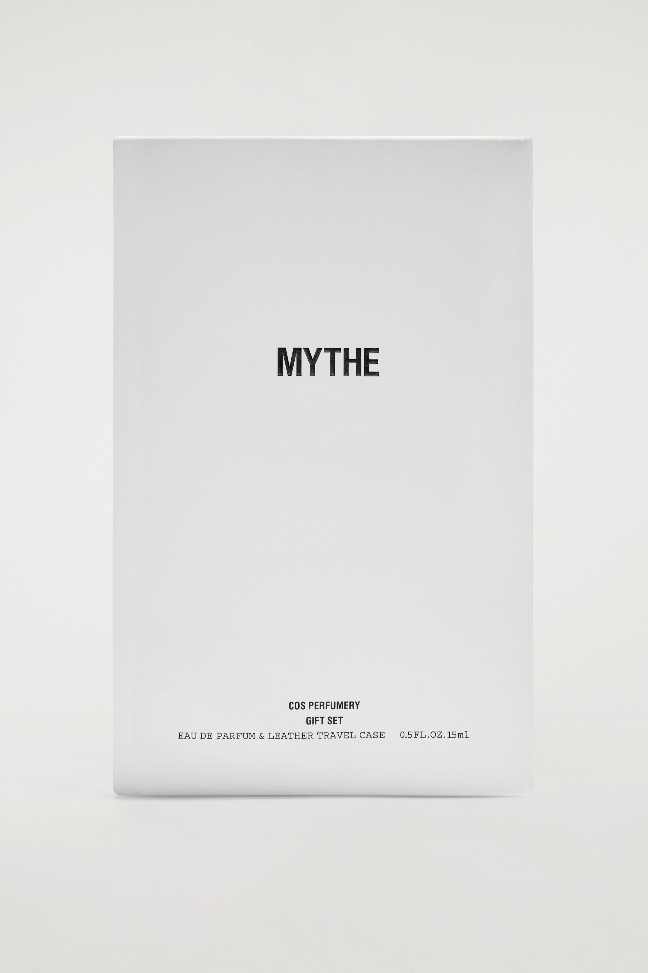 A white rectangular gift box stands upright, featuring a minimalist front-facing design. The centre displays the word "MYTHE" in bold black lettering, while smaller black text near the bottom details the product as a "COS PERFUMERY GIFT SET" containing an "EAU DE PARFUM & LEATHER TRAVEL CASE." A white rectangular gift box stands upright, featuring a minimalist front-facing design. The centre displays the word "MYTHE" in bold black lettering, while smaller black text near the bottom details the product as a "COS PERFUMERY GIFT SET" containing an "EAU DE PARFUM & LEATHER TRAVEL CASE."