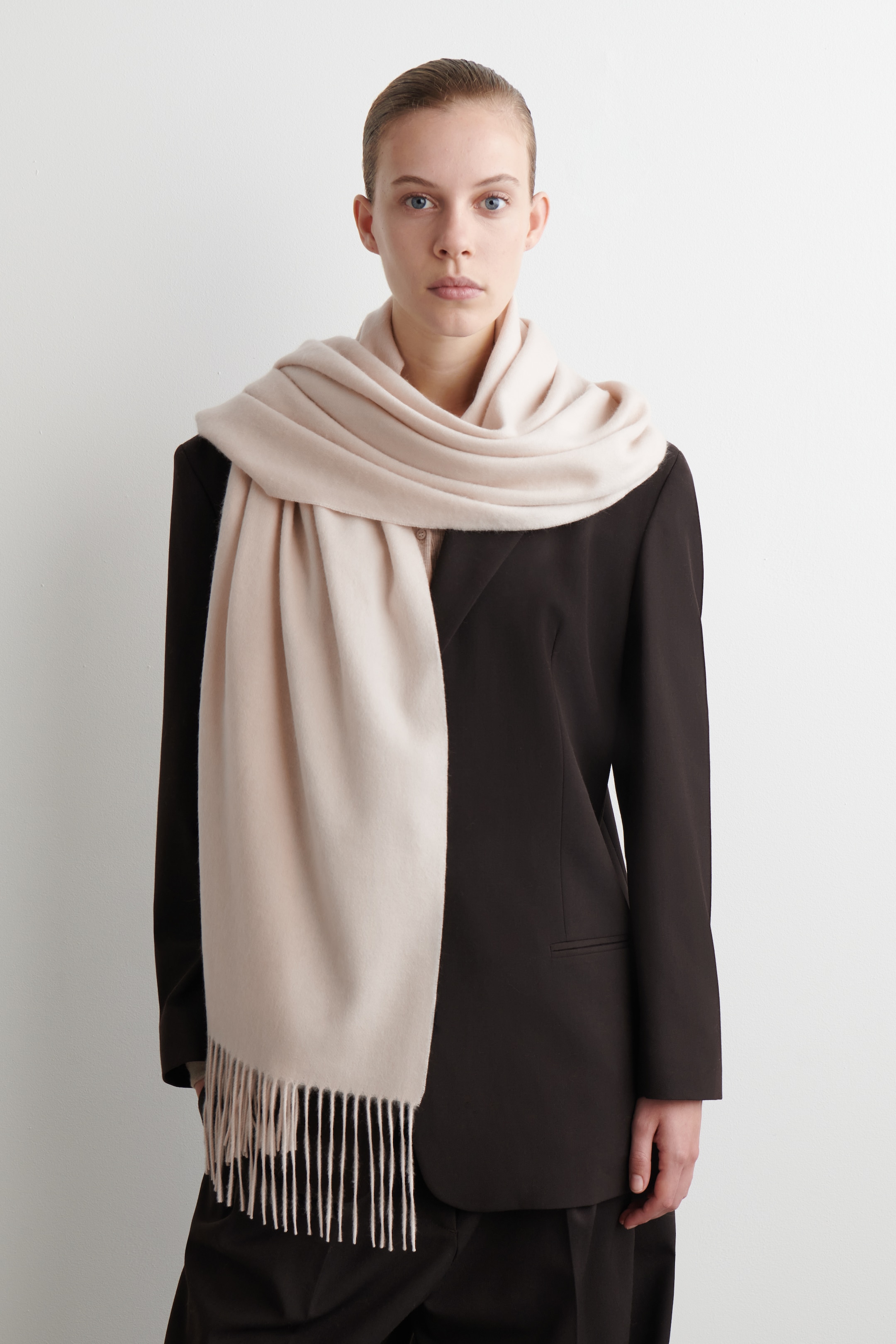 Agrandir l'image: Une écharpe beige pâle est drapée autour du cou et sur les épaules, avec une extrémité tombant sur le devant. Cette écharpe à la texture lisse présente de fines franges le long de son bord inférieur et est portée sur un blazer foncé.