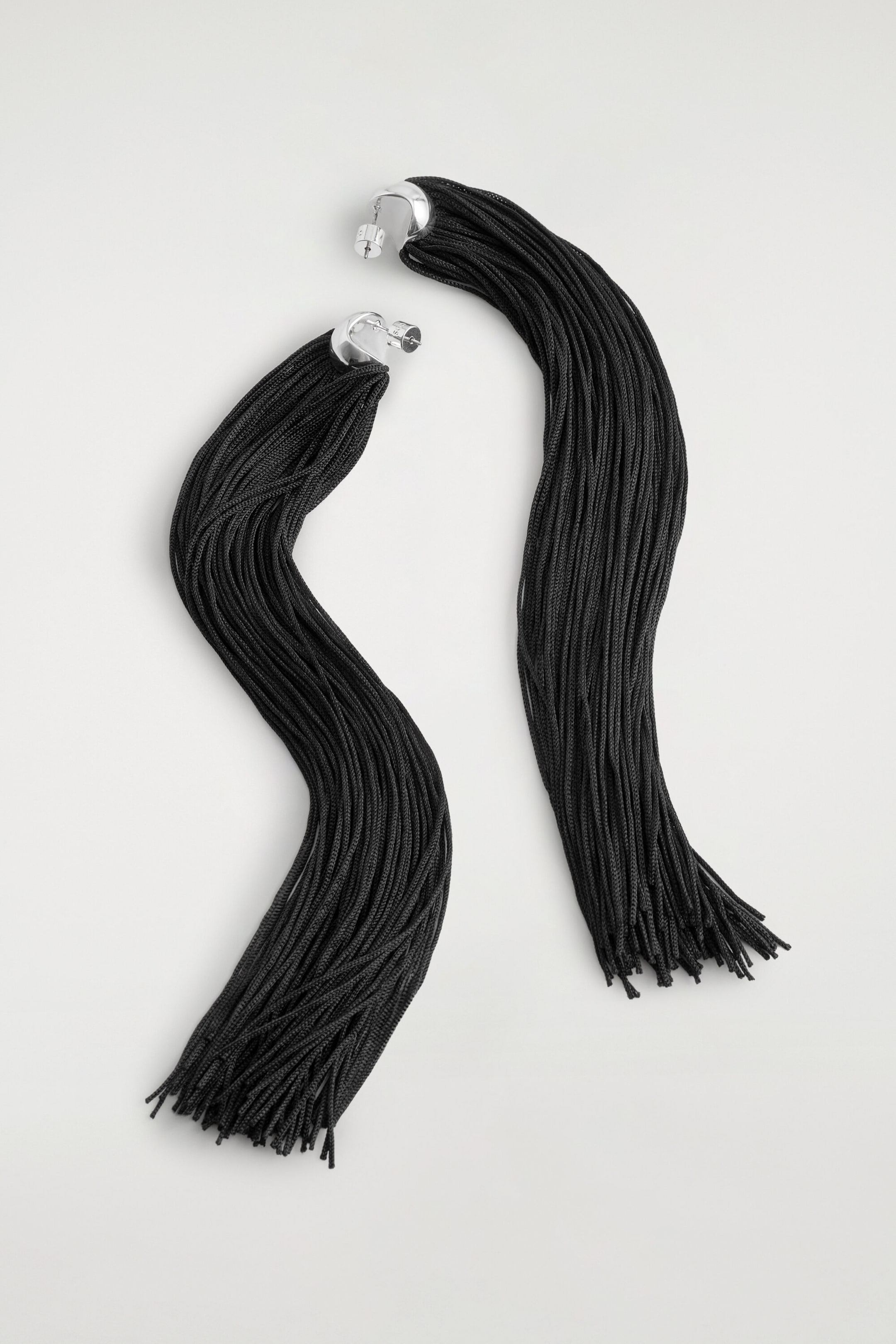 Fringed earrings are shown in a flat lay, featuring curved, silver-tone metal studs with post-back closures. Long, dense bundles of black cord fringe extend from the metal bases, draping downward in soft, wavy curves.