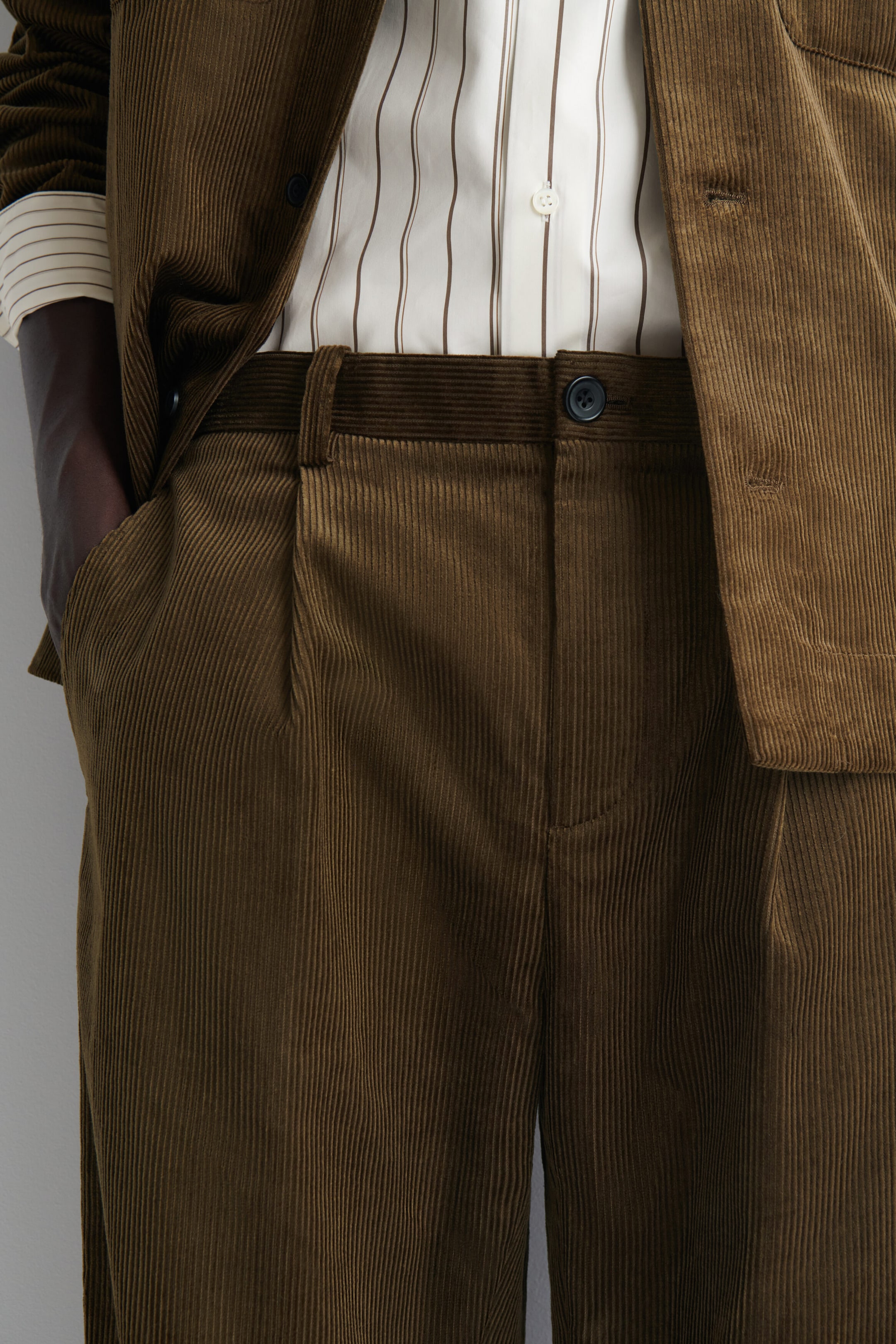 The Pleated Relaxed-Fit Tapered Corduroy Pants in a dark olive-brown shade are shown from the front, featuring a single pleat on each side, belt loops, and a dark button closure. They showcase a relaxed fit with a hand slipped into the side pocket, paired with a tucked-in striped shirt and a matching open corduroy blazer. The Pleated Relaxed-Fit Tapered Corduroy Pants in a dark olive-brown shade are shown from the front, featuring a single pleat on each side, belt loops, and a dark button closure. They showcase a relaxed fit with a hand slipped into the side pocket, paired with a tucked-in striped shirt and a matching open corduroy blazer.